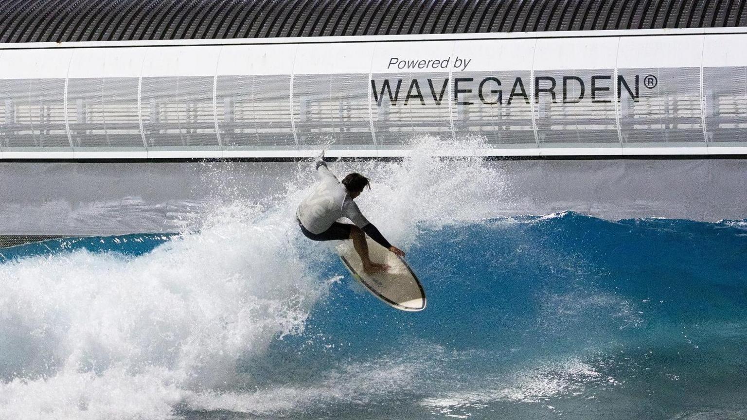 A man on a surfboard rides a wave at The Wave in Bristol. The water is a blue-green colour with white foam, and he is wearing a white top and black shorts