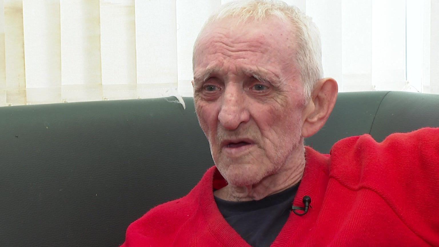 An elderly, white male with short white hair dressed in a red jumper sits on a sofa and looks at the camera