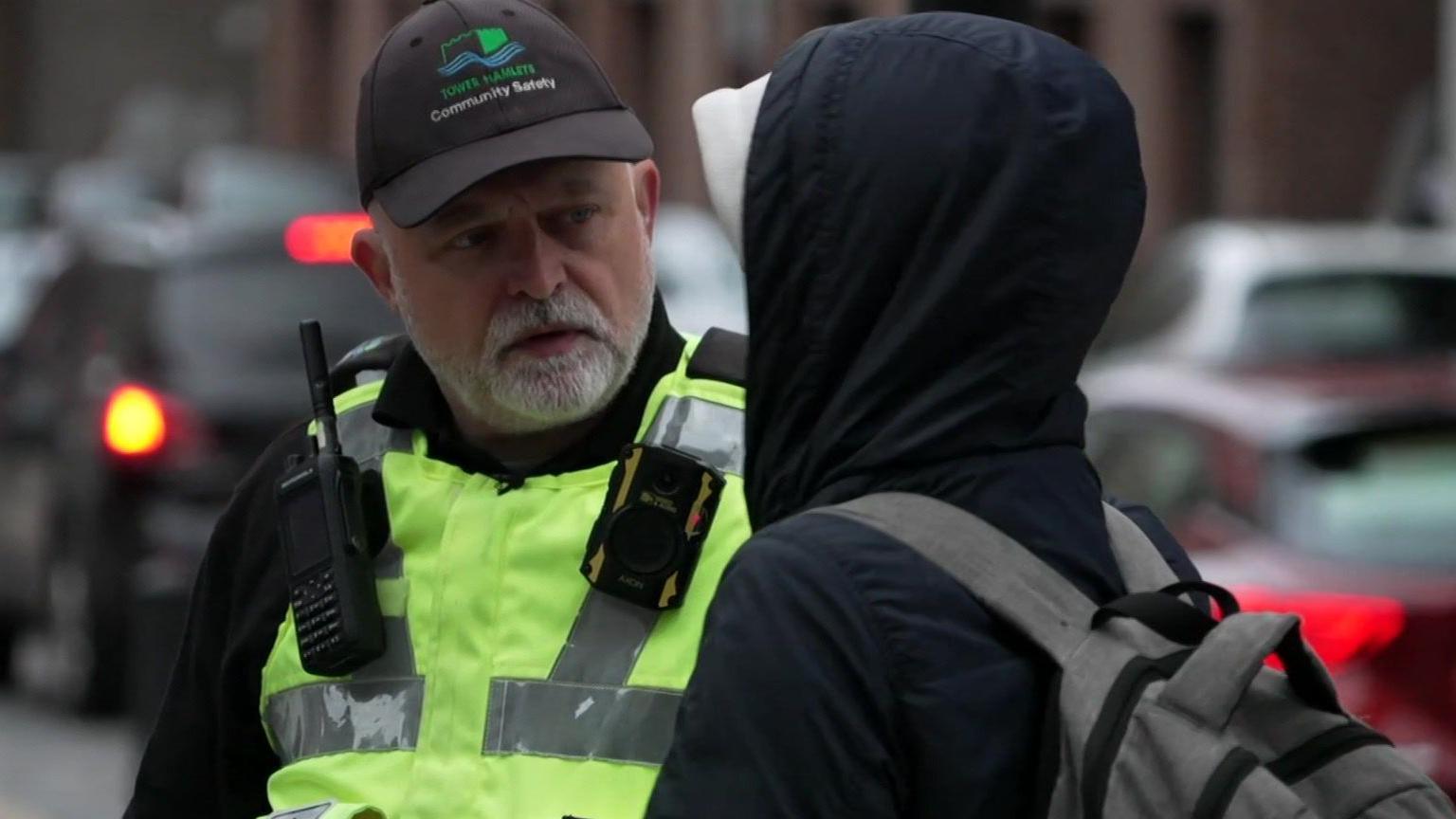 John Fish wears a hi-vis jacket with a radio and body worn camera on the front. He has a baseball cap on his head bearing the council logo. He is on a street, talking to a man with his back to the camera, and the hood of his coat up