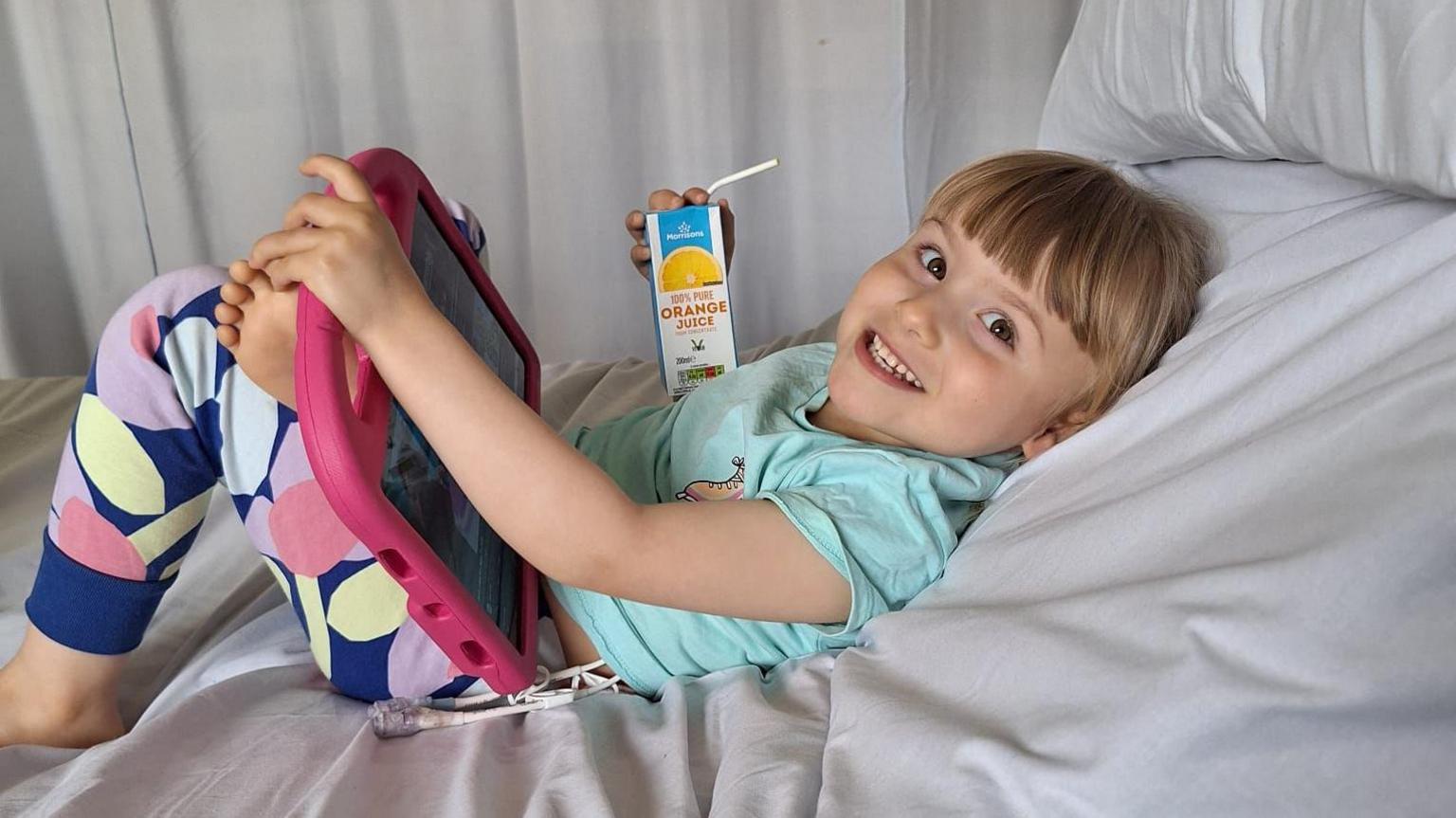 Freya Dixon lying n a hospital bed smiling and watching an tablet drinking a carton of juice and wearing a turquoise tshirt and brightly patterned leggings 