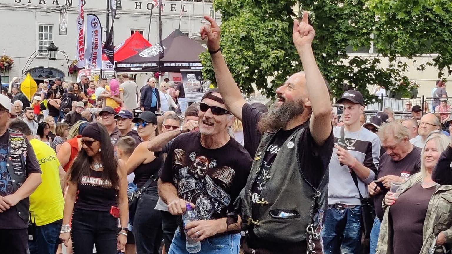 A large crowd of people at a bike meet in Calne. Two men are at the forefront of the picture. One has his arms up in the arm while the other is holding a bottle of water. Both are smiling
