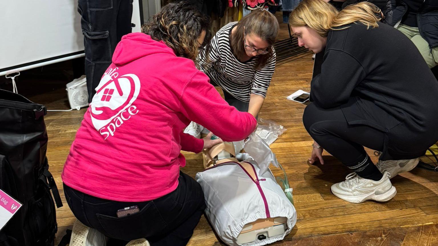 A group of women learning CPR on a dummy. They are all crouched on the floor around the dummy. One woman is wearing a bright pink hoodie with a Safe Space logo printed on the back. 