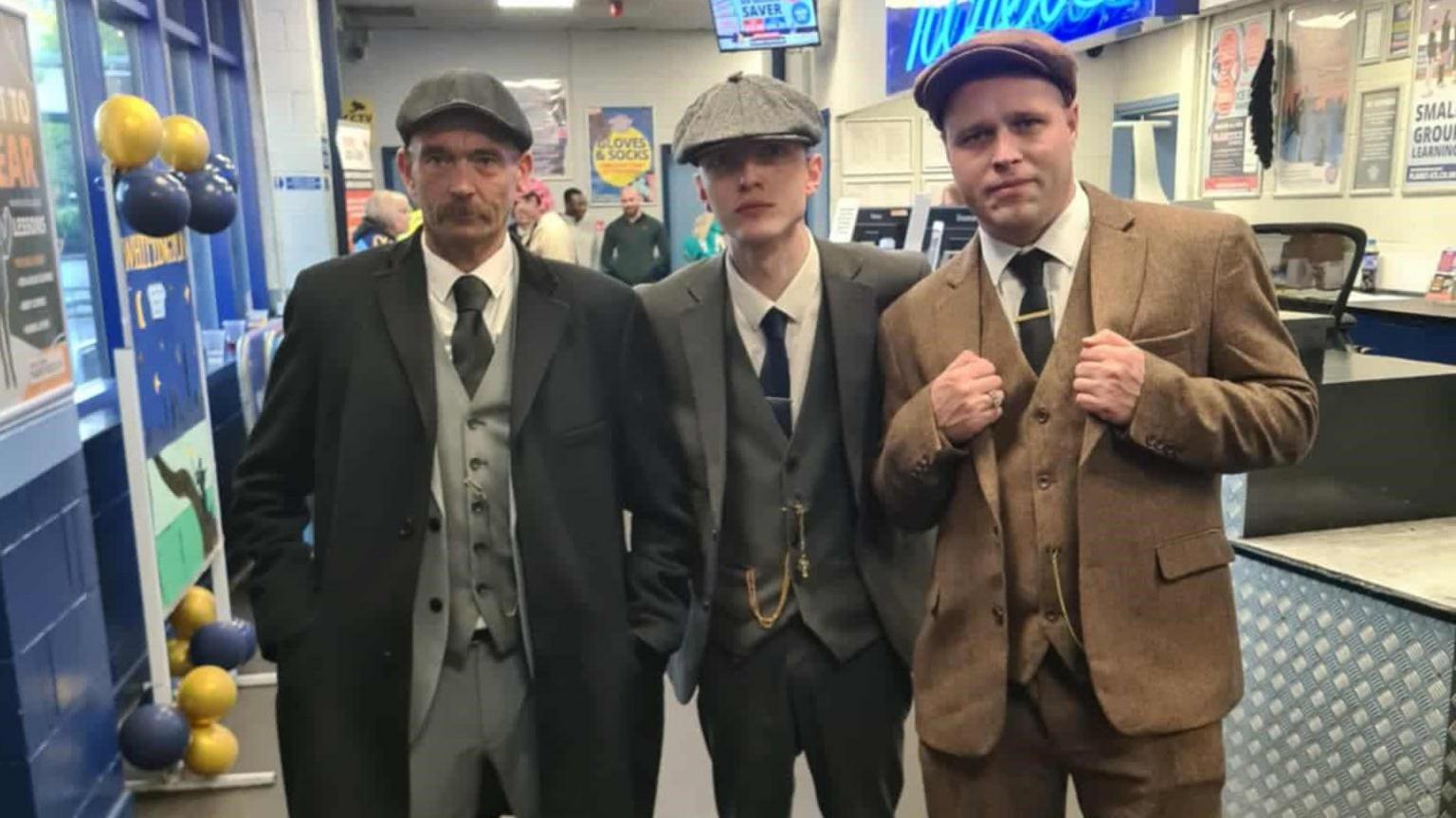 Three men wearing flat caps and tweed suits stand together in a shop. One man is holding his fists together in front of him.
