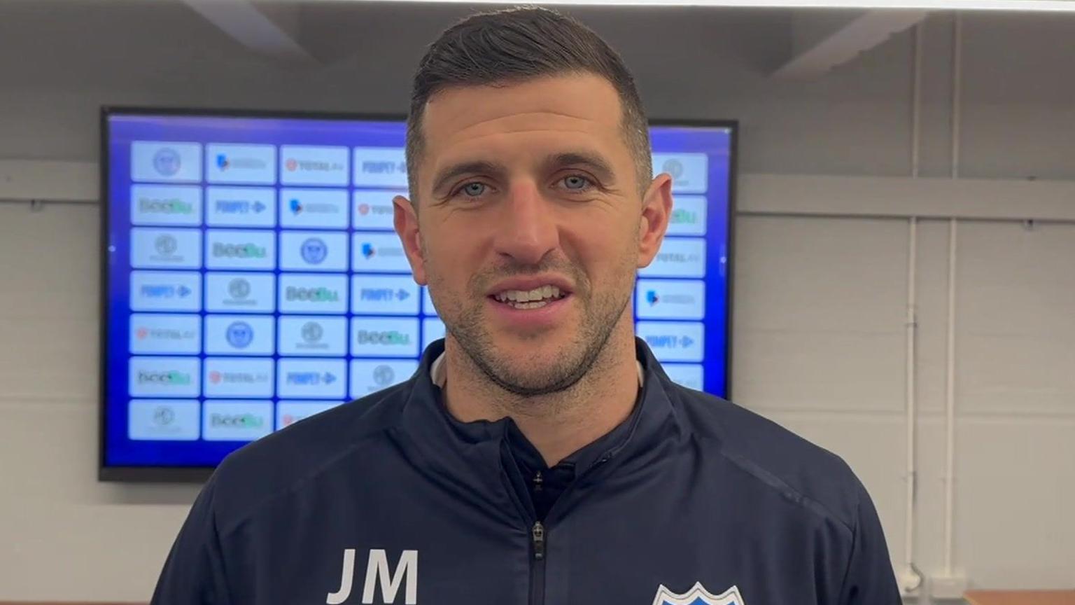 John Mousinho smiling at the camera. He has dark hair, blue eyes and is wearing a navy tracksuit top with the initials JM on the left hand side.