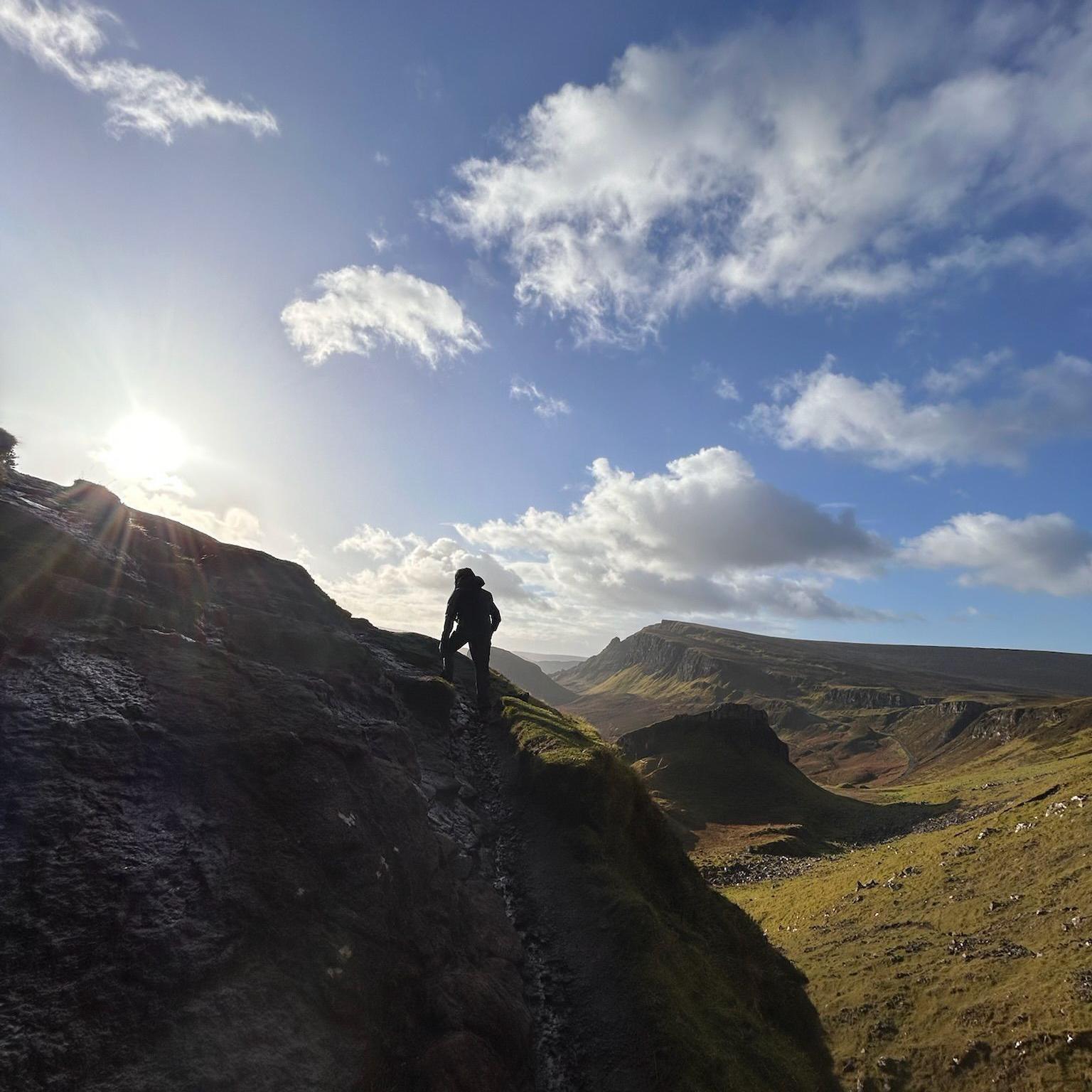 The silhouette of a man can be seen on a mountain coming in from the left side of the picture. There is blue sky above it with a few clouds. The sun is also in the top left of the picture. In the background, there are green fields and dark hills.