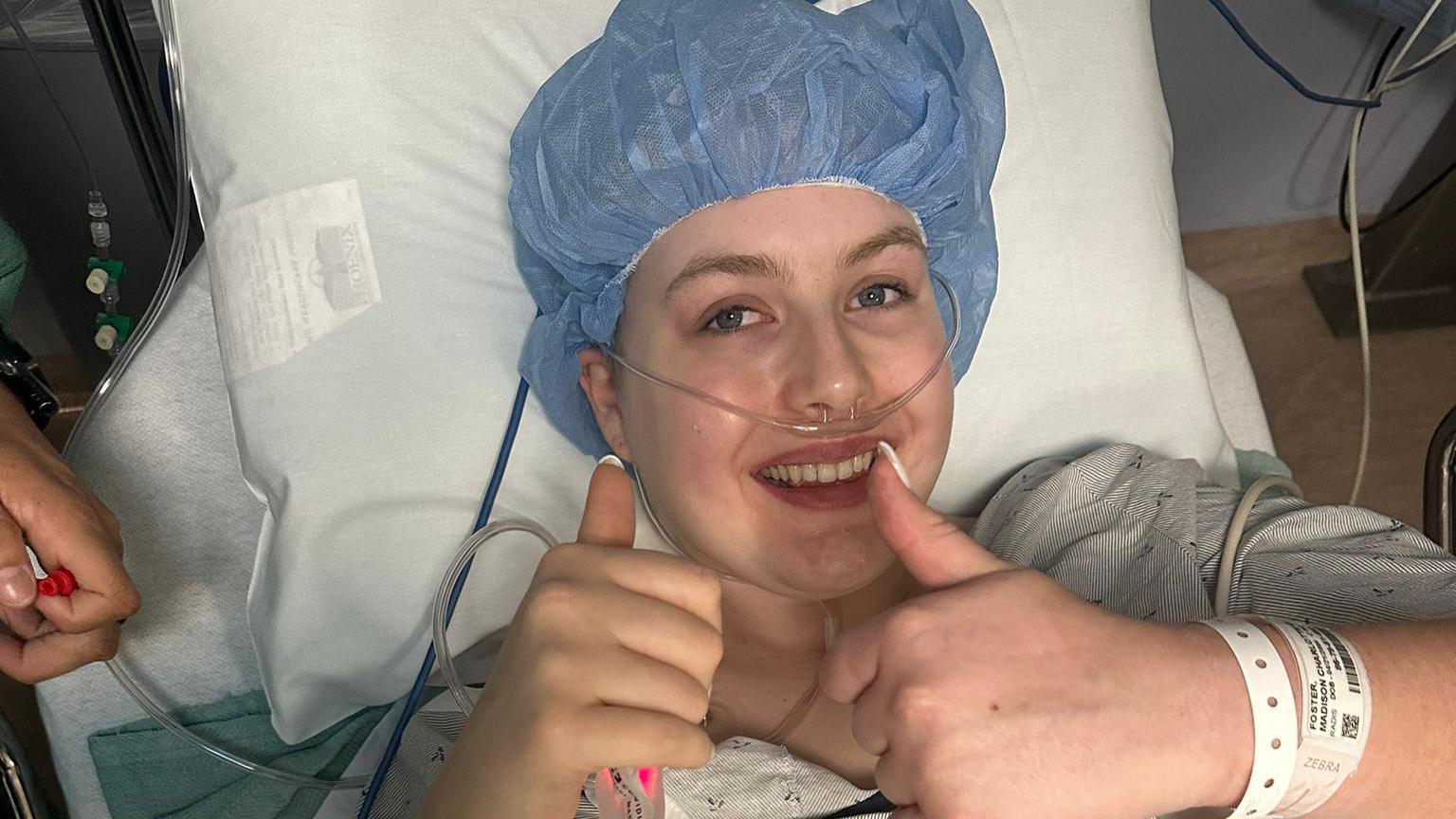 A woman wearing a blue disposable hair net lying on a hospital bed with tubes and wires attached. She is smiling and holding two thumbs up to the camera.