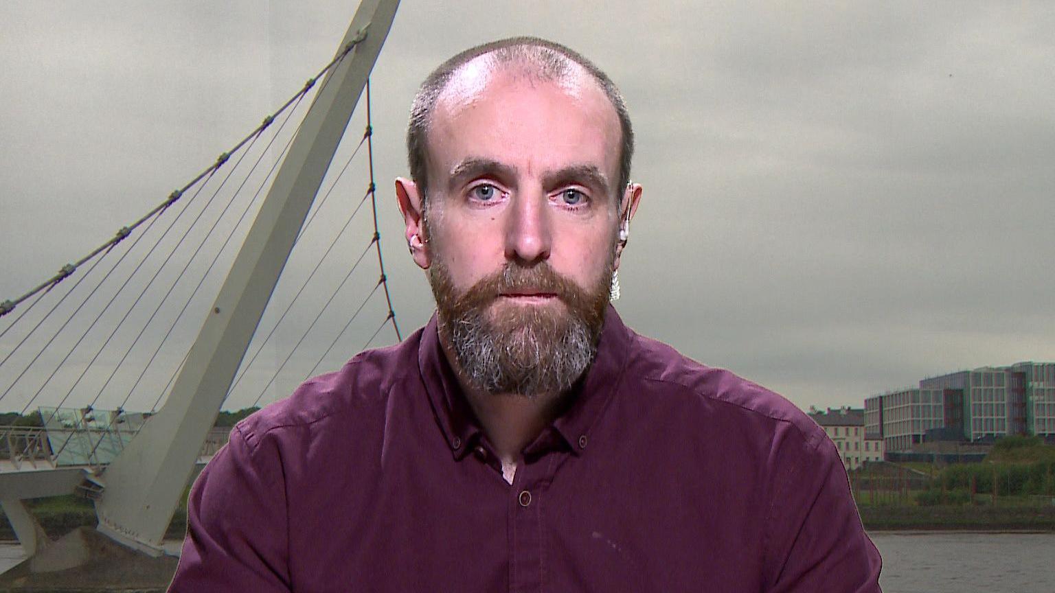 Mark Durkan - a man with receding hairline and a brown beard streaked with grey. He is wearing a plum coloured shirt and is sitting against a background of the peace bridge in Londonderry.