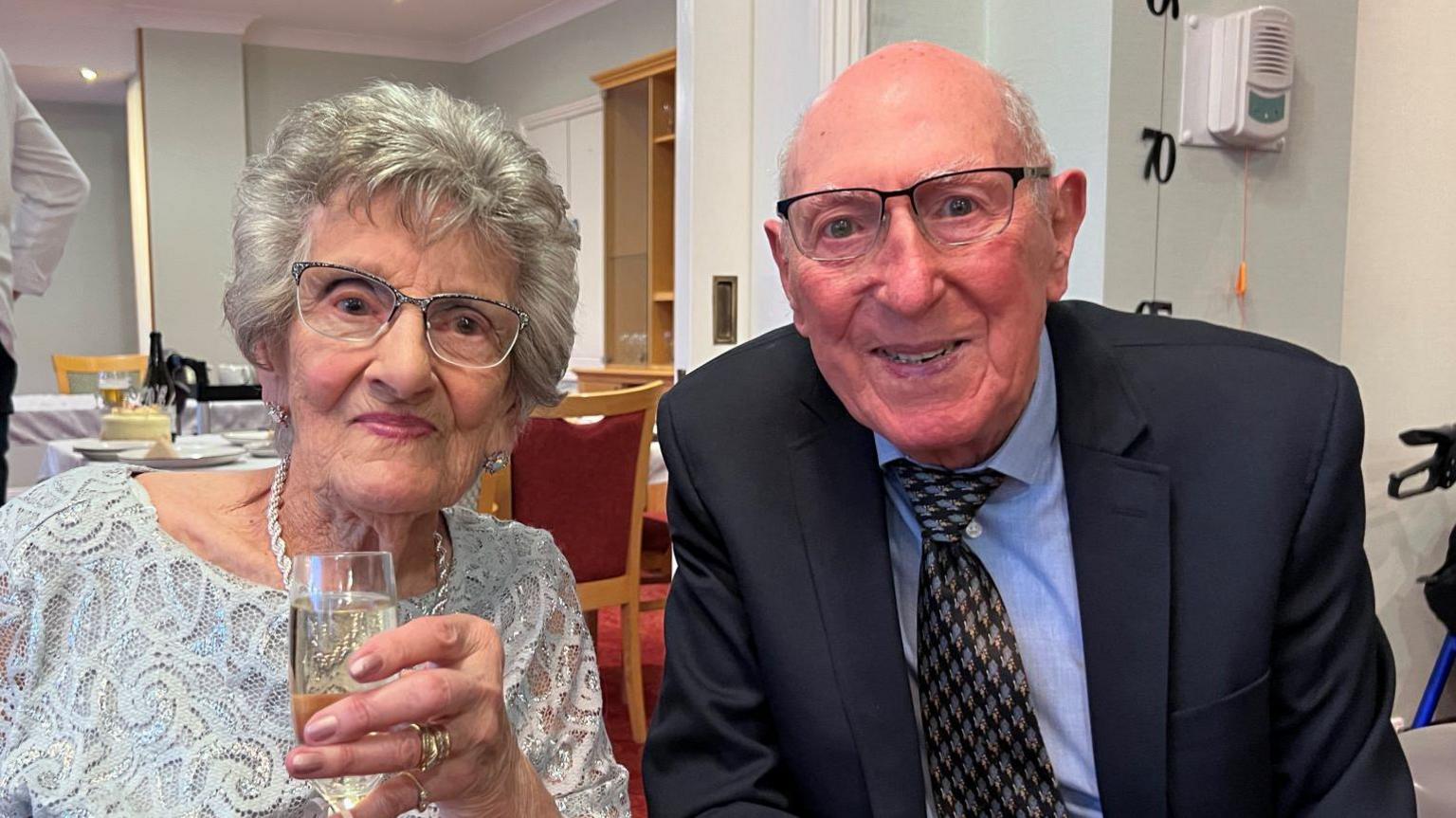 Nancy Ashpool holding a glass of prosecco while sitting next to Ron Ashpool and wearing a silver dress. Mr Ashpool is wearing a suit and tie. Both are smiling and sitting in a living room during the anniversary party.