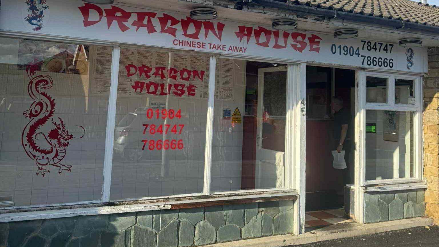 A Chinese takeaway called Dragon House which has been cleaned after being covered in graffiti. It has large windows with red writing on and a picture of a dragon.
