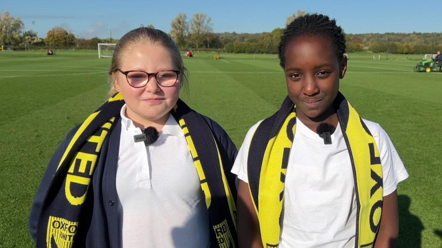 Two Year 6 students wearing white s-shirts and yellow Oxford United scarves stand in front of the football pitch smiling at the camera.