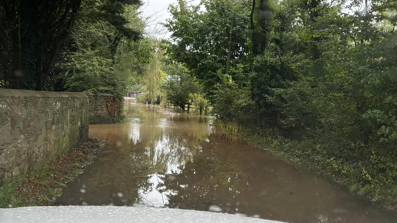 Floodwater across a country road, with greenery on either side of the road and an old stone wall to the left. The picture is taken from the front seat of a car which cannot pass. 