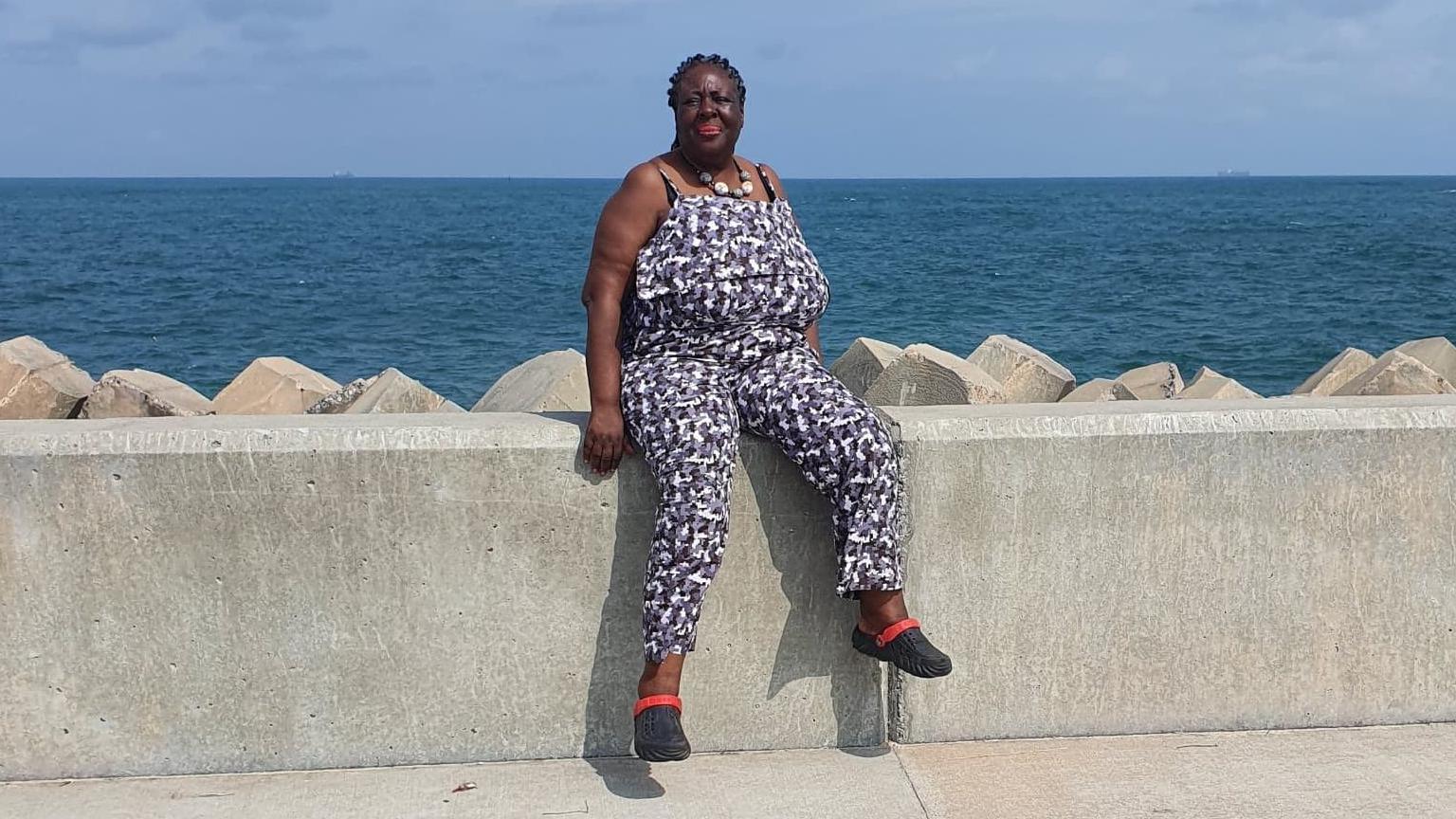 A woman in a pattern jumpsuit and sandals sits on a wall infront of the ocean, posing and smiling for the picture