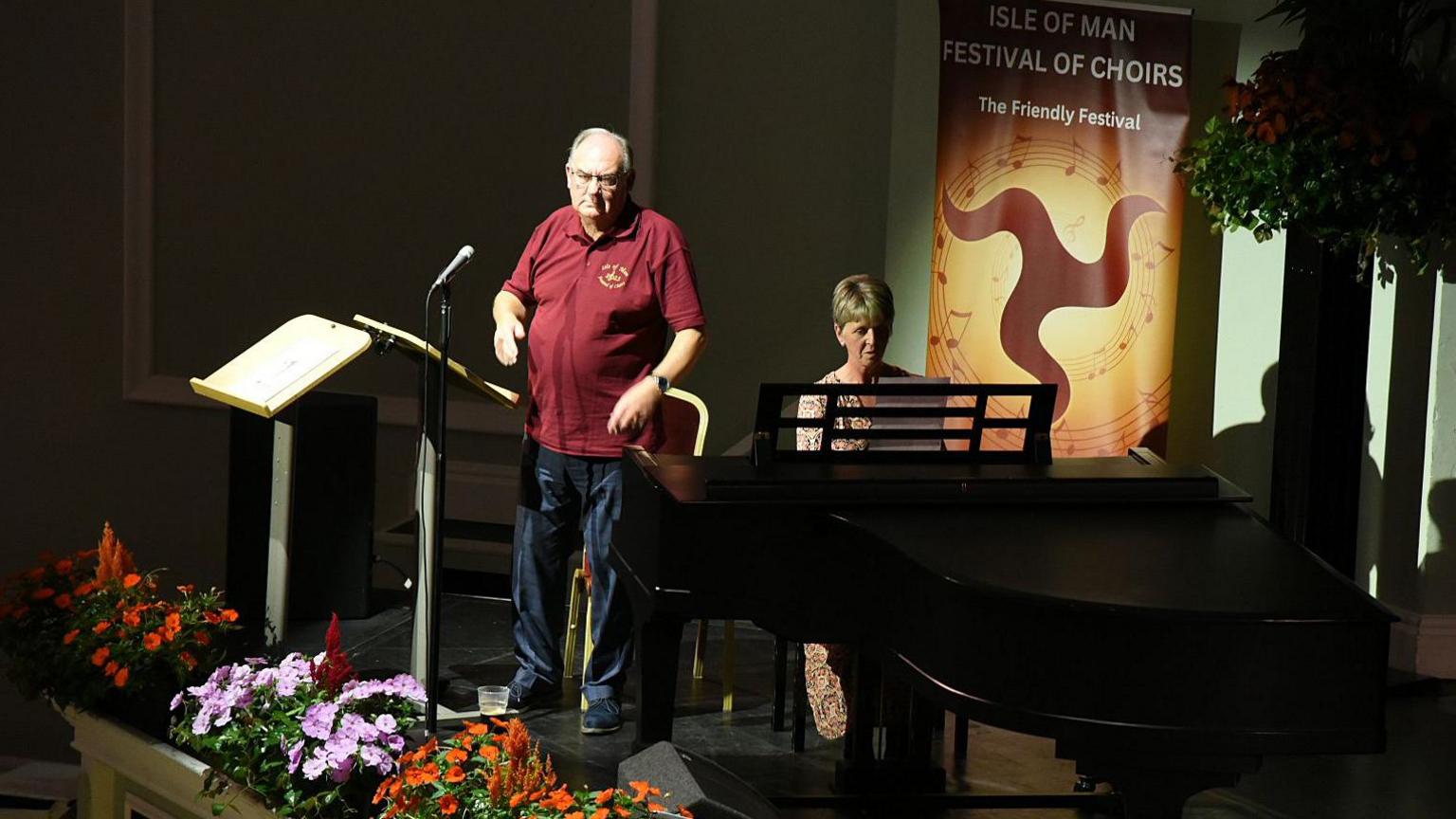 Geoff Corkish, a man with grey hair and glasses wears a red T-shirt. He is on a stage with a woman who is at a piano next to him and there is a sign in the background that reads Isle of Man Festival of Choirs.