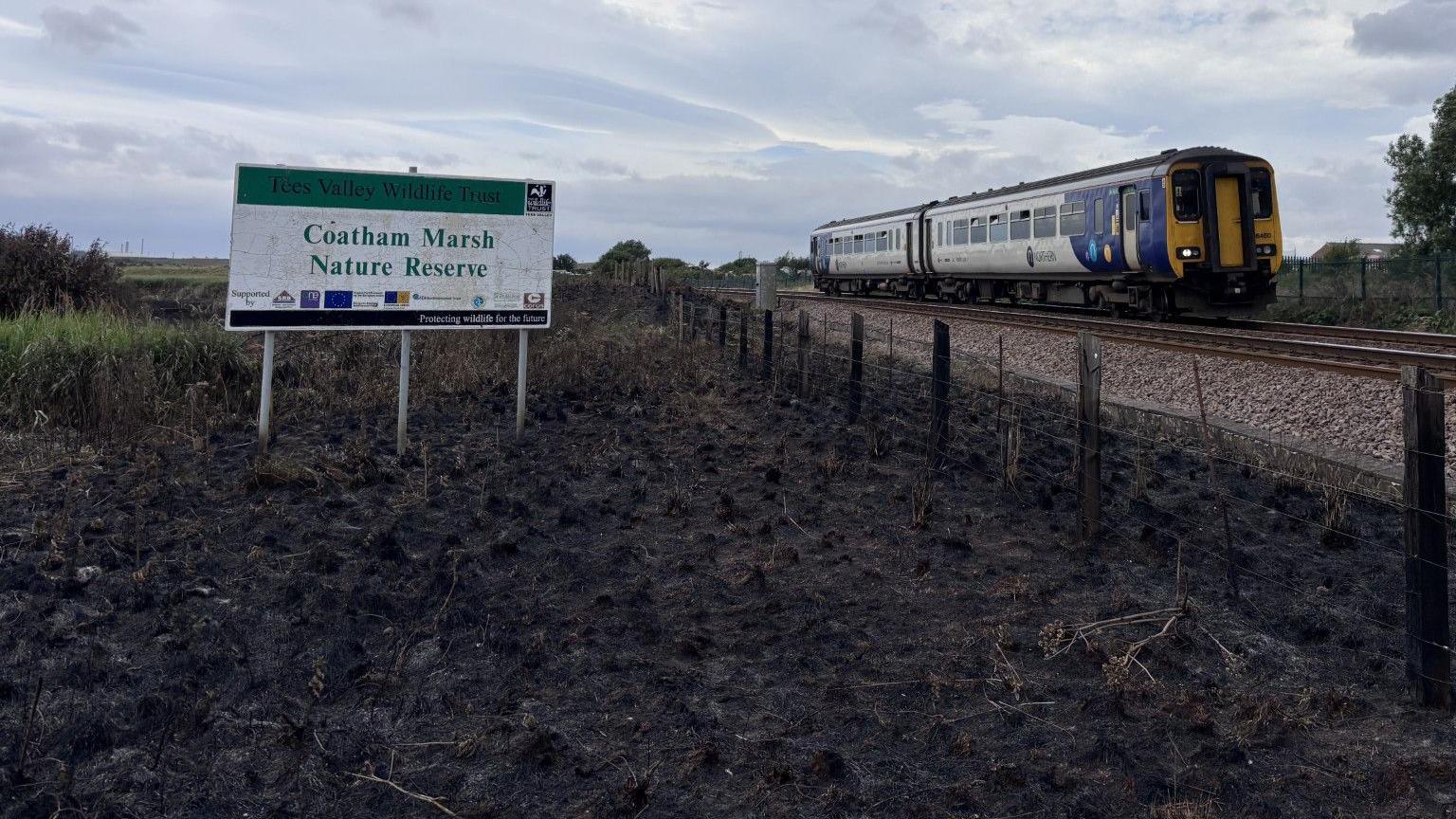 A train is passing close to a burned field with a sign reading 'Coatham Marsh Nature Reserve' standing in it.