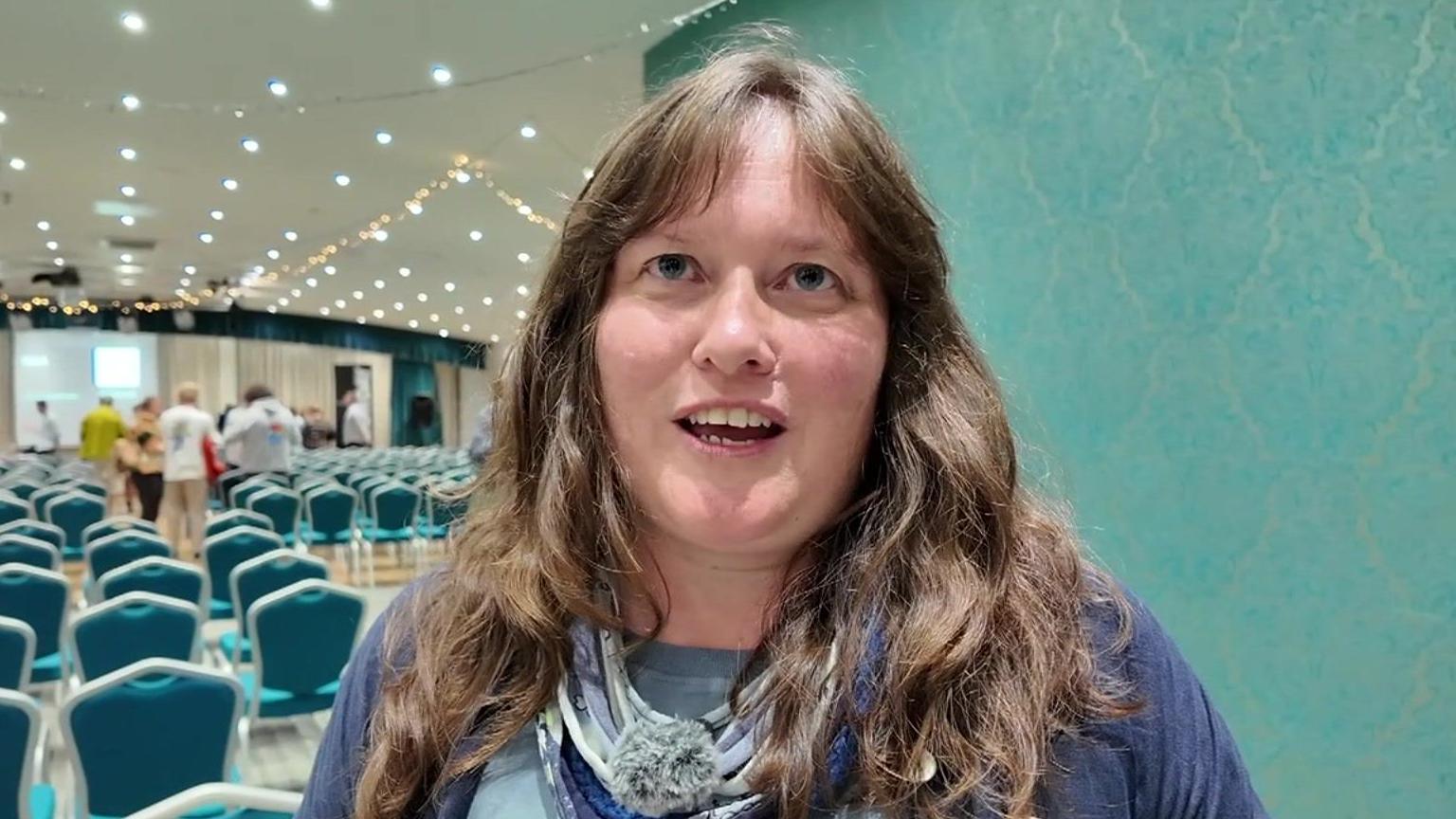 A woman with long brown hair and her mouth open while talking and looking up towards a camera inside a room filled with blue chairs following a meeting. She is wearing a purple top and is stood in front of a turquoise wall.