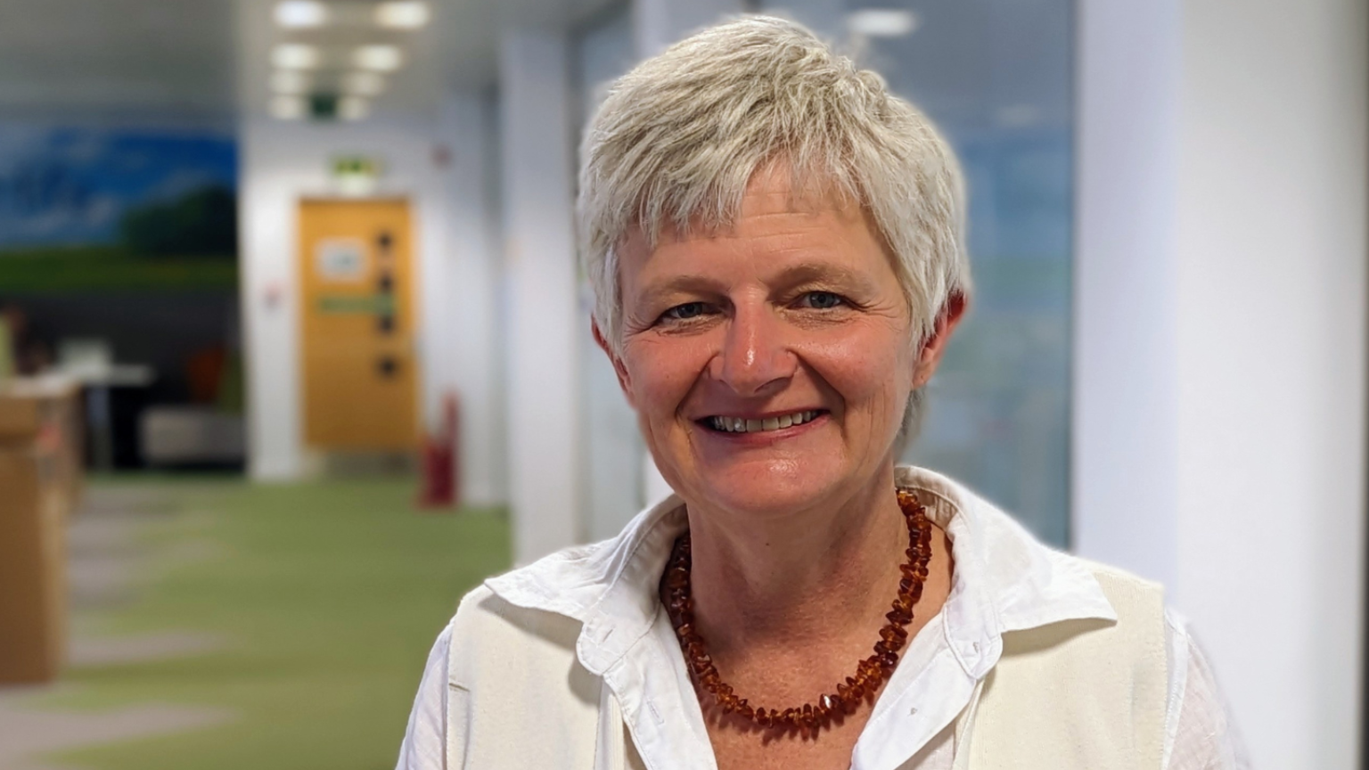 Katherine Crawford, a smart lady with short white hair smiles in her office which has scenic murals on the walls. She wears a white shirt and a red beaded necklace.
