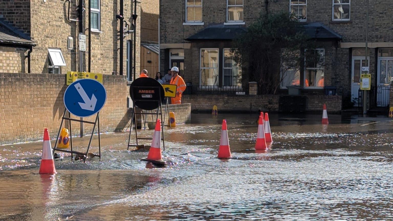 Water gushing out of an apparent hole on a road in Oxford. There are cones and signs around the hole.