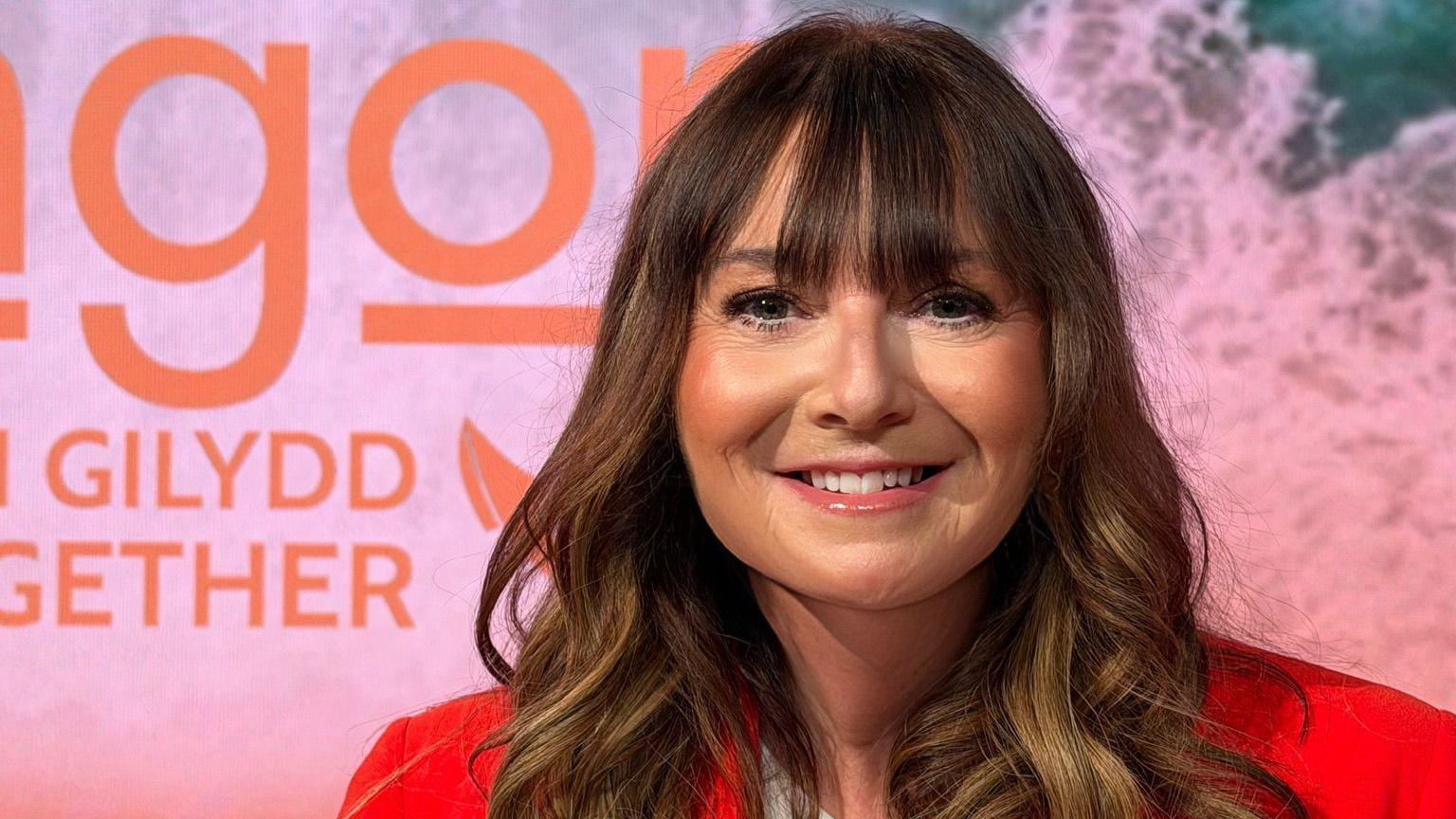 Jo-Anne gyda'i gwallt wedi'i chyrlio ar set teledu. Mae hi'n gwenu gyda siaced coch llachar a crys-t gwyn Angor. Mae logo Angor i'w weld ar y sgrin tu ol hi. 