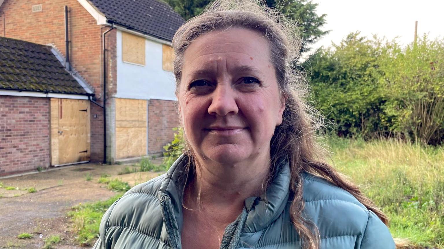 Lucy Nethsingha looking direct to camera. She is outside. To the left of the image and over her shoulder is a boarded-up, detached, brick-built house. On the right of the image is long grass and some bushes, with a taller tree visible in the background. Lucy is wearing a puffa-style, seagreen colour coat, with her light brown hair coming over one shoulder.