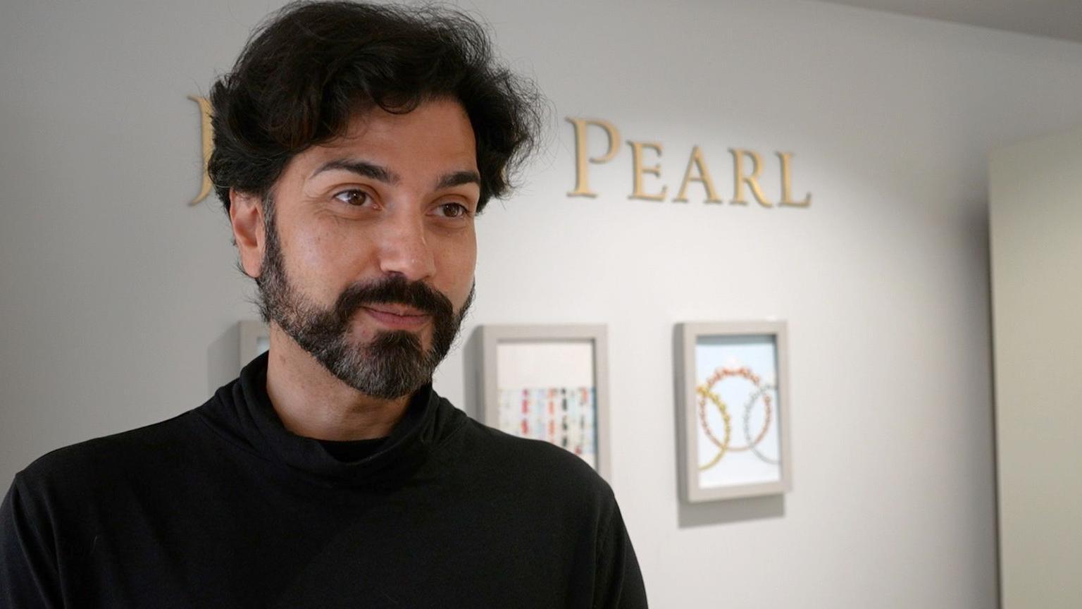 A man dressed in all back looking to the side of the camera with a wall with two frames and the word Pearl in it.