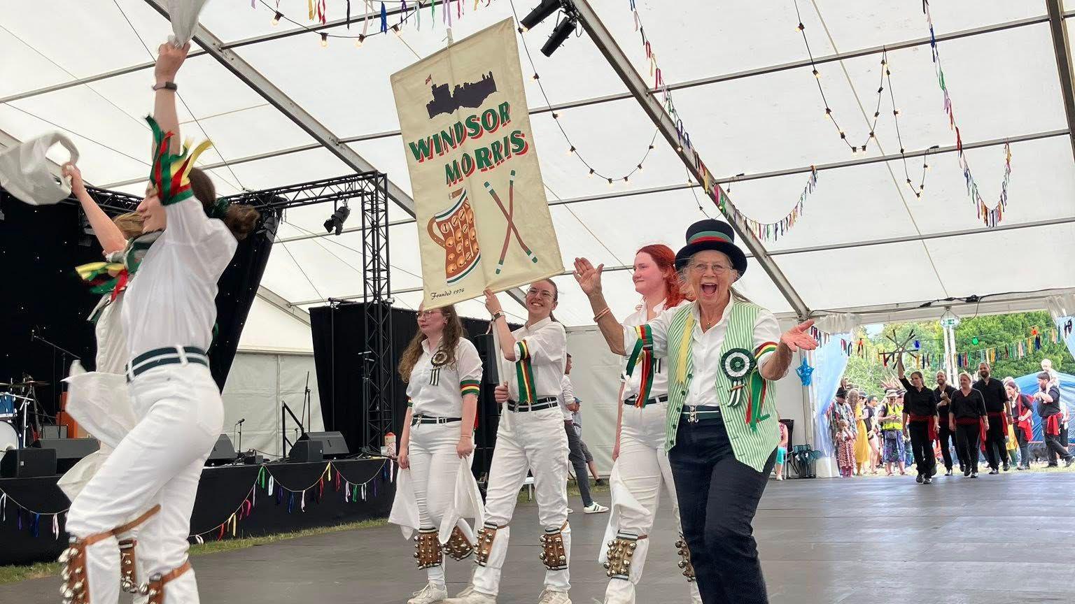 A parade of morris dancers making their way through a marquee. Most of the women are wearing white tops and trousers with bells on. One woman closest to the camera is wearing black jeans and a green and white striped waistcoat, she is holding her hands up, in a jazz-hands style, and wearing a black top hat.