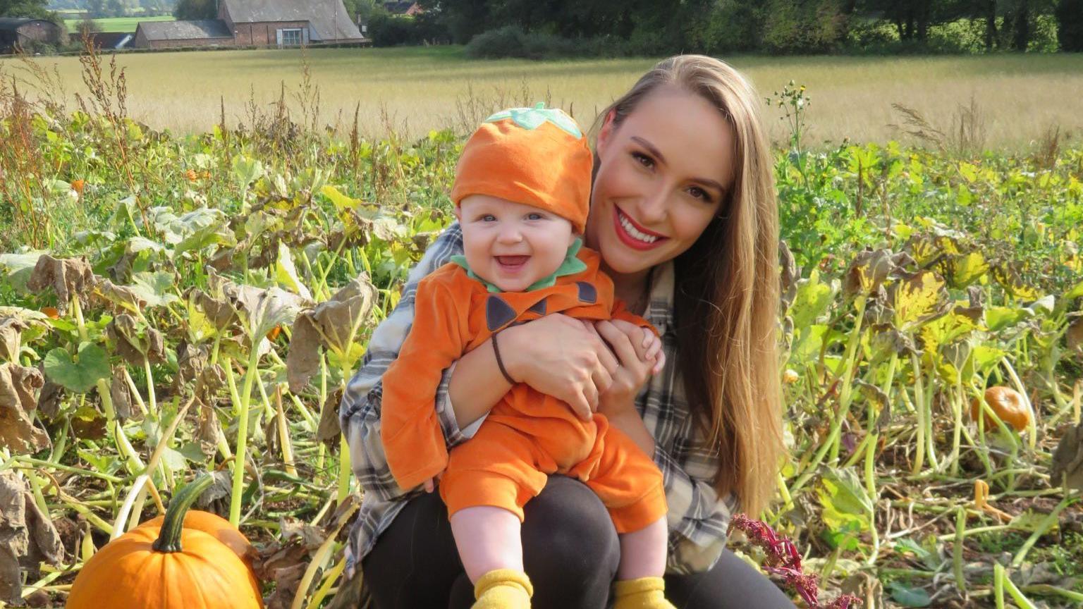 Emma and Leo in a pumpkin patch . Leo has an orange jumper and hat and is sitting on mum's knee with a big smile