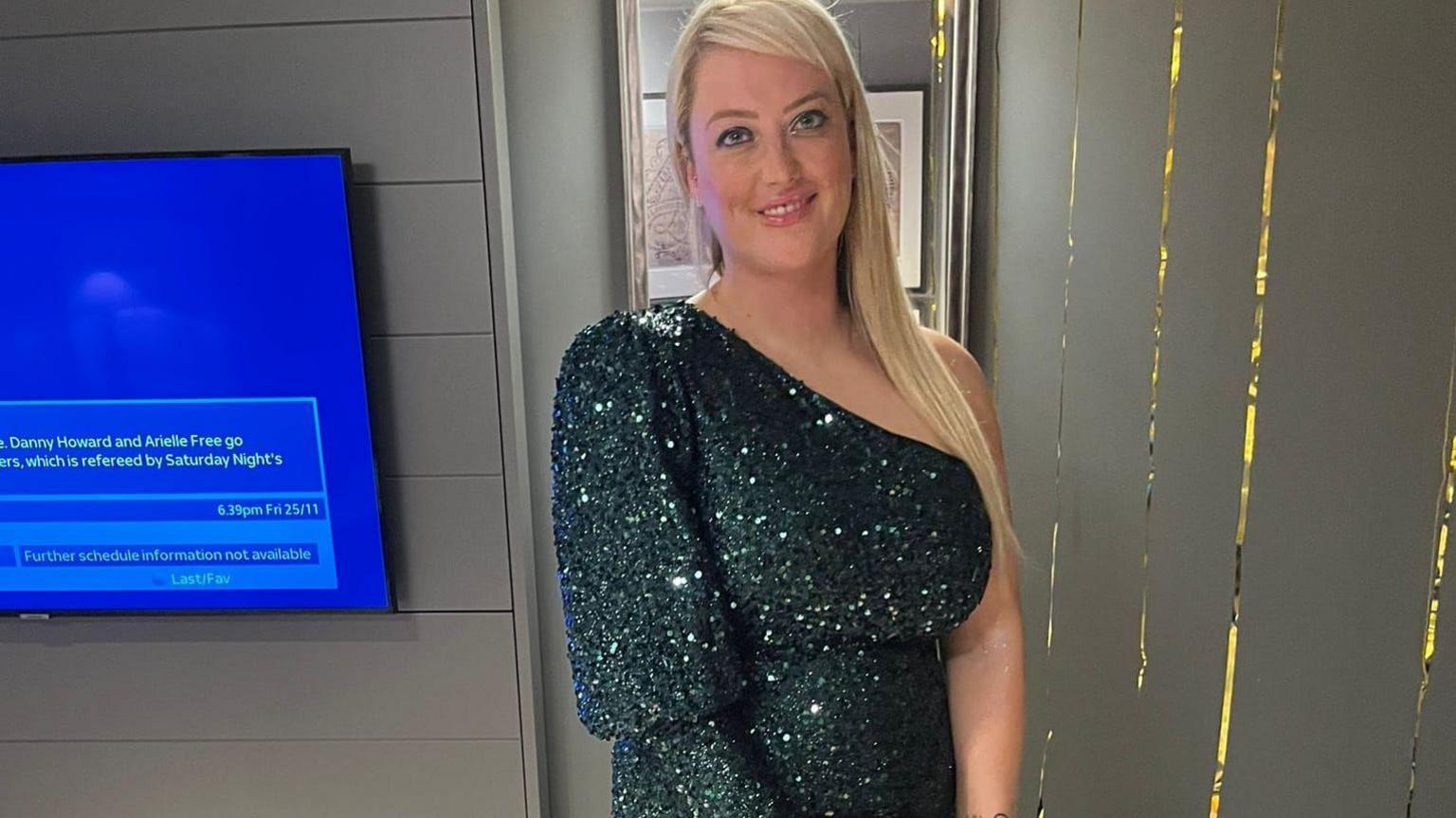 Blonde woman standing in front of a TV wearing an off the shoulder green sparkly dress