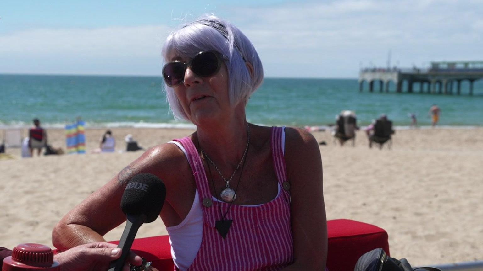 Denise is wearing a pink and white striped pinafore top and is sitting on a red sofa in front of the beach