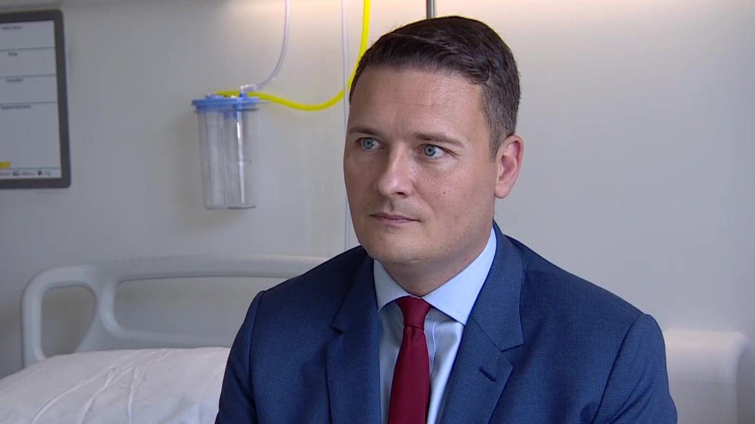 Wes Streeting in a blue suit and red tie sat next to a hospital bed