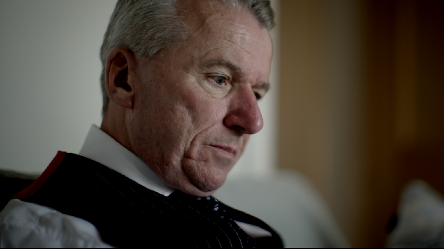 A man wearing a white shirt, black waistcoat and black tie looks down at some paperwork in an office setting