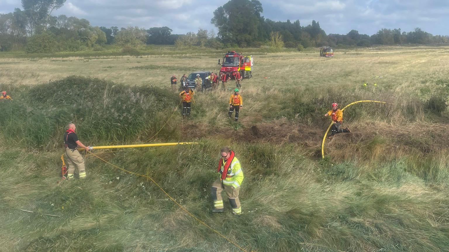 Firefighters, a car and a specialist fire service all-terrain vehicle on farmland in Suffolk. The grass is thick and long, and it is cloudy.