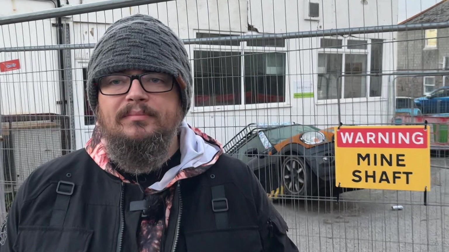 A man with a beard and a black beanie hat looks at the camera - behind him is his car in the mineshaft