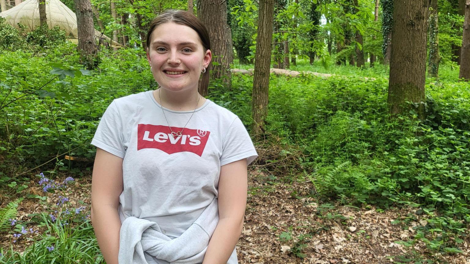 Eva Fillingham wearing a white Levi's t shirt and hoop earrings, smiling at the camera. She has a white hoody tied around her waist and there are trees and woodland behind her.