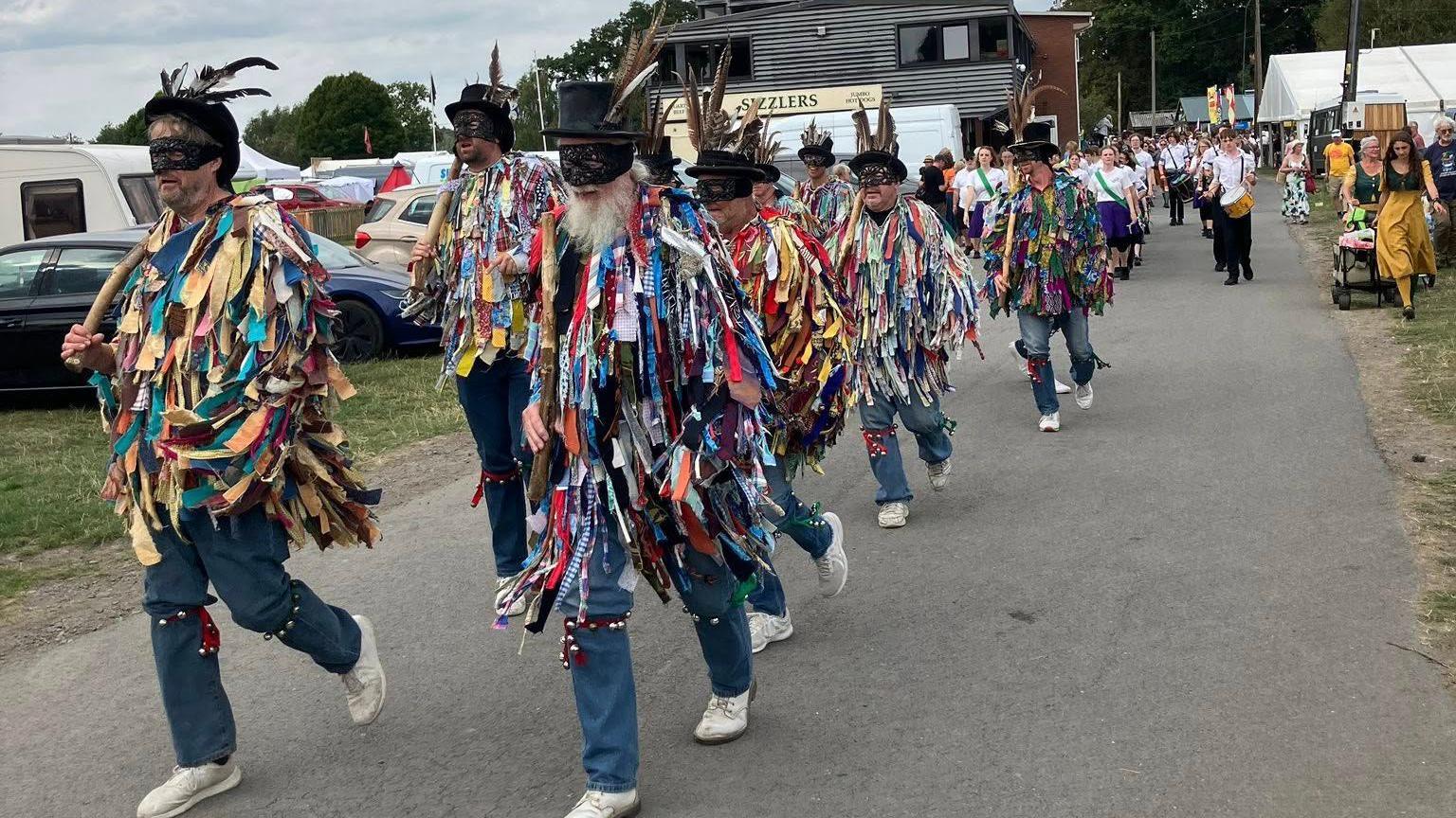 Men walking side by side, as part of a Morris parade dressed in tunics made from strips of cloth over their normal clothes. They also have bells strapped just above their knees and are making their way through a park. 