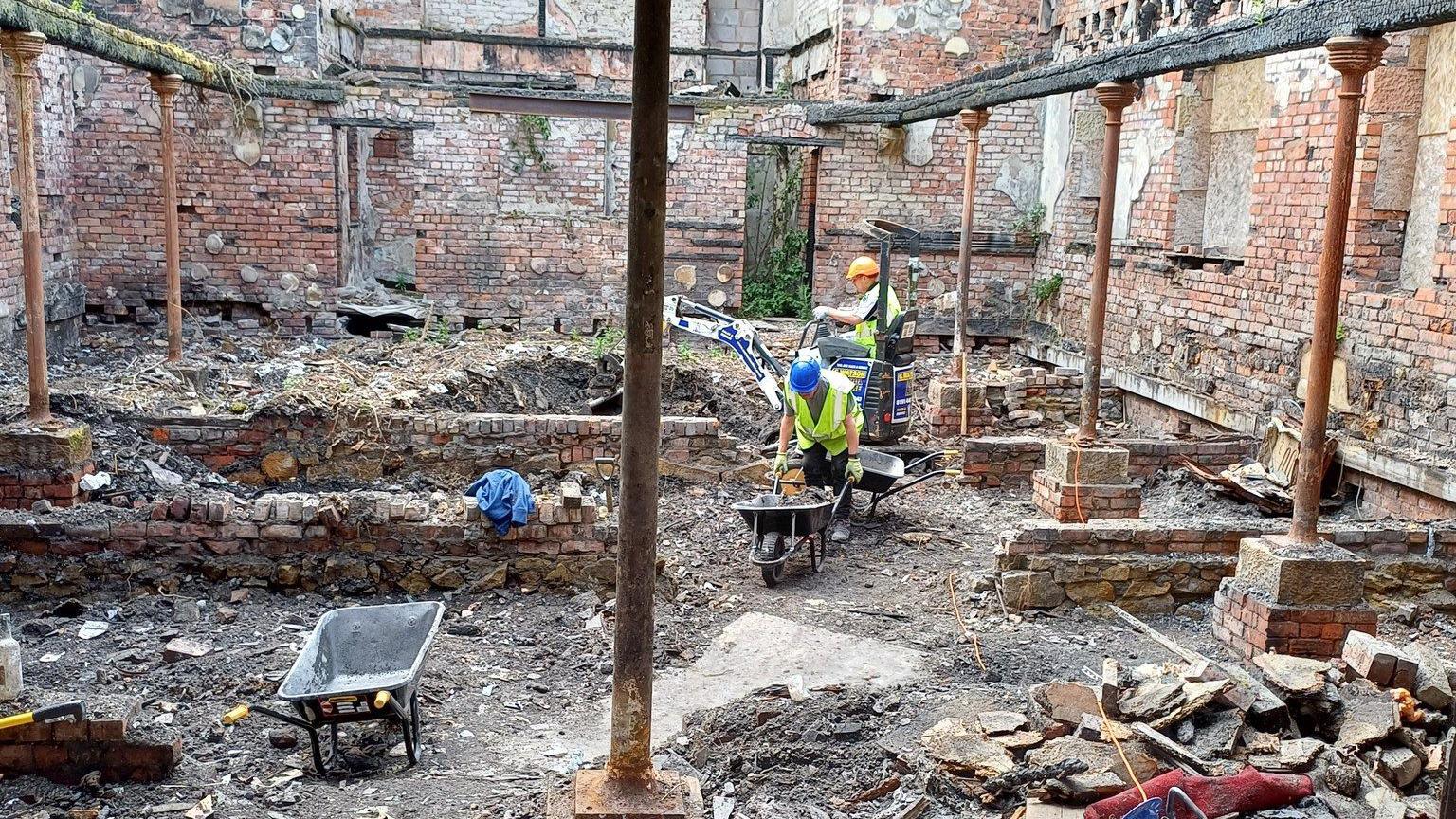 Two people wearing hard hats and hi-vis jackets clear rubble from the derelict church site. One is pushing a wheelbarrow and the other is working a small digger. The ground is uneven and metal props are holding up some stonework.