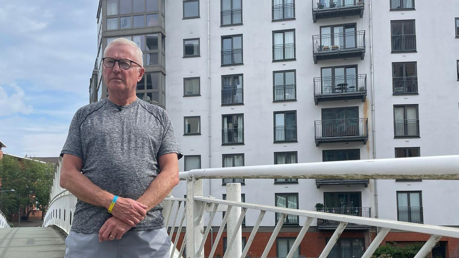 A man in a grey t-shirt and trousers stands on a bridge in front of a white tower block, with several black balconies visible