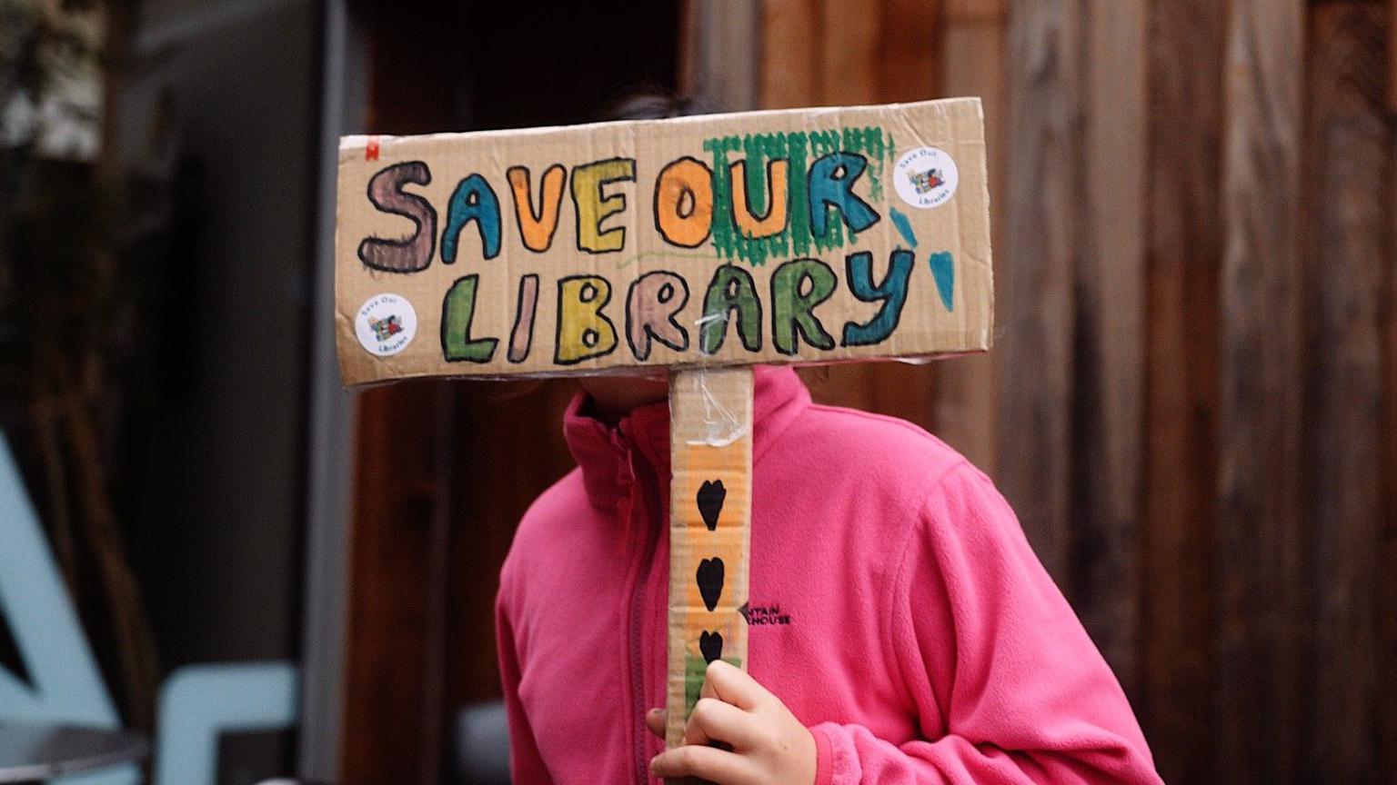 A girl wearing a pink hoodie is holding a sign reading 'Save Our Library'.