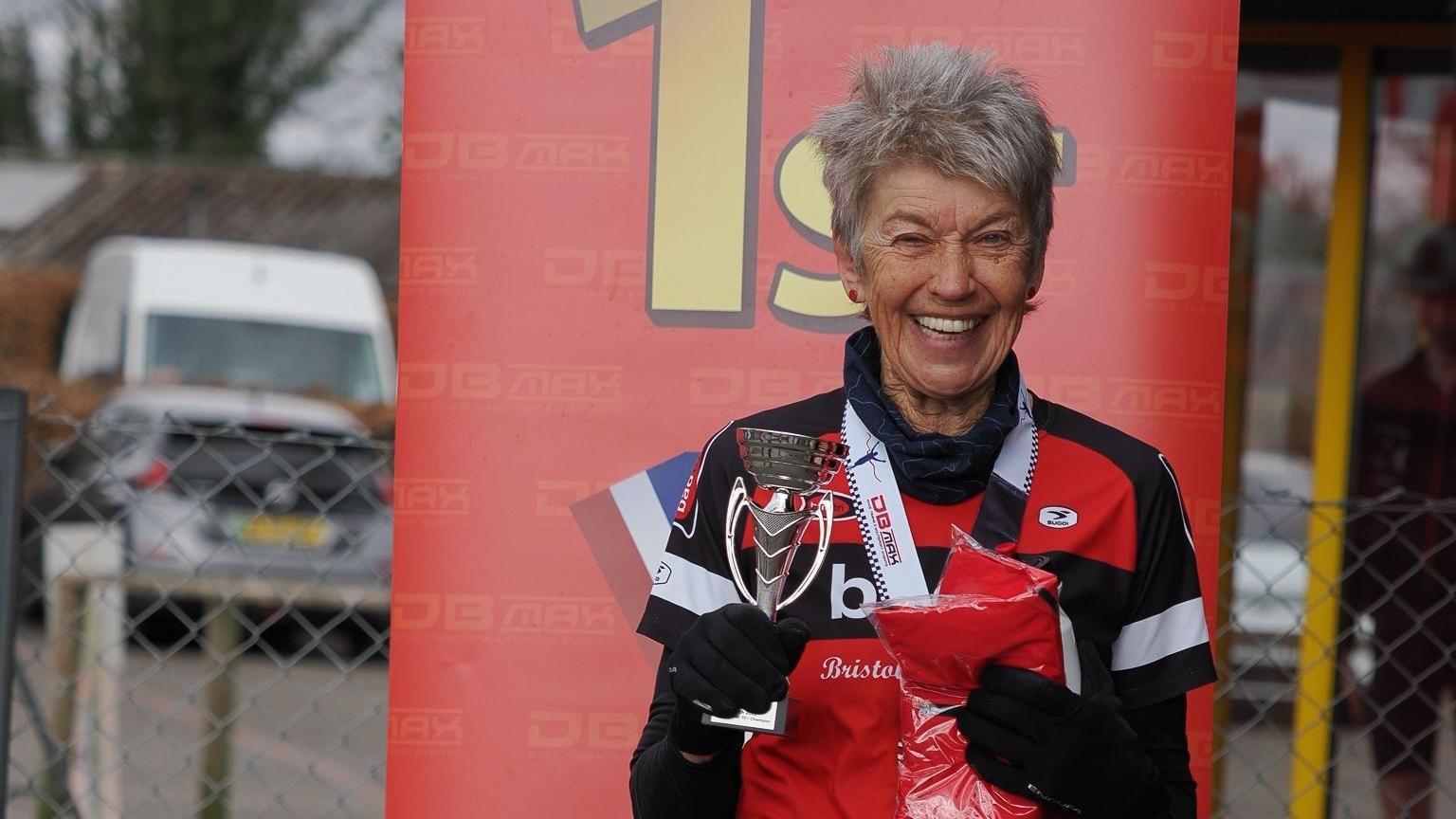 Anne Dockery wearing red, black and white sports gear. She smiles at the camera as she holds a silver trophy in one hand and a red finisher t-shirt in the other. She has a medal with a white ribbon around her neck and is standing in front of a red 1st place banner.