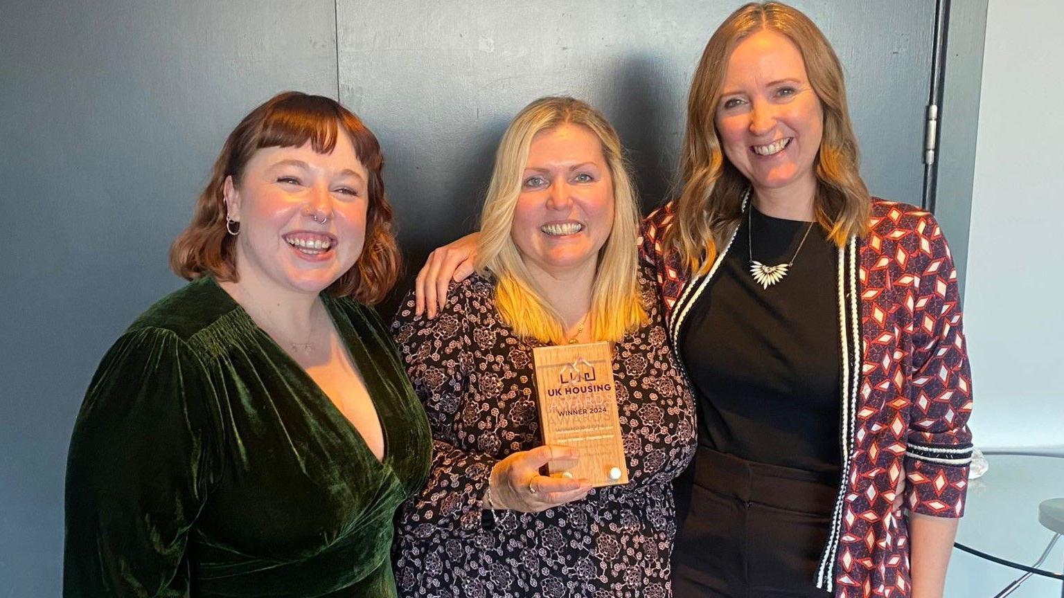 Three women standing next to each other. The lady in the middle is holding an award. 
