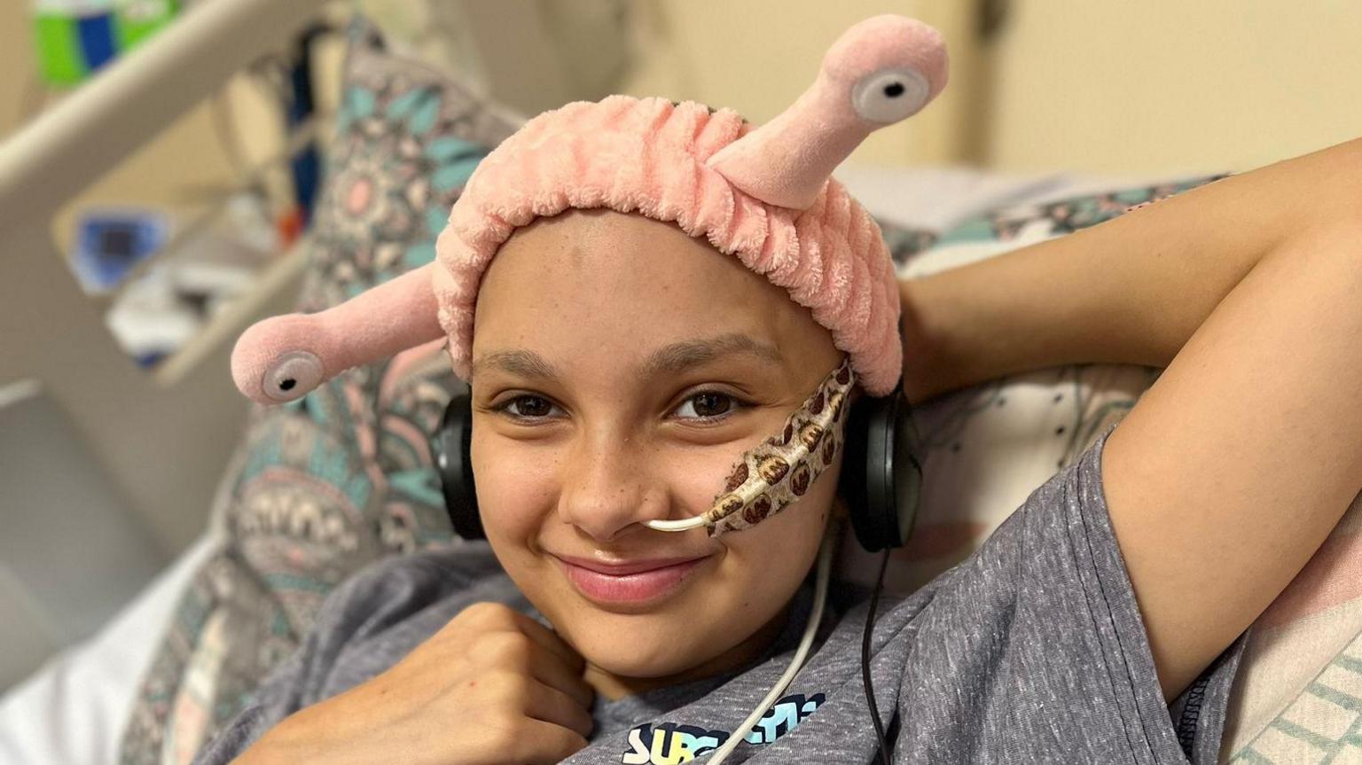 12-year-old Amelie is lying in bed in hospital, she has a tube in her nose and a plaster on, and is wearing a pink headband.