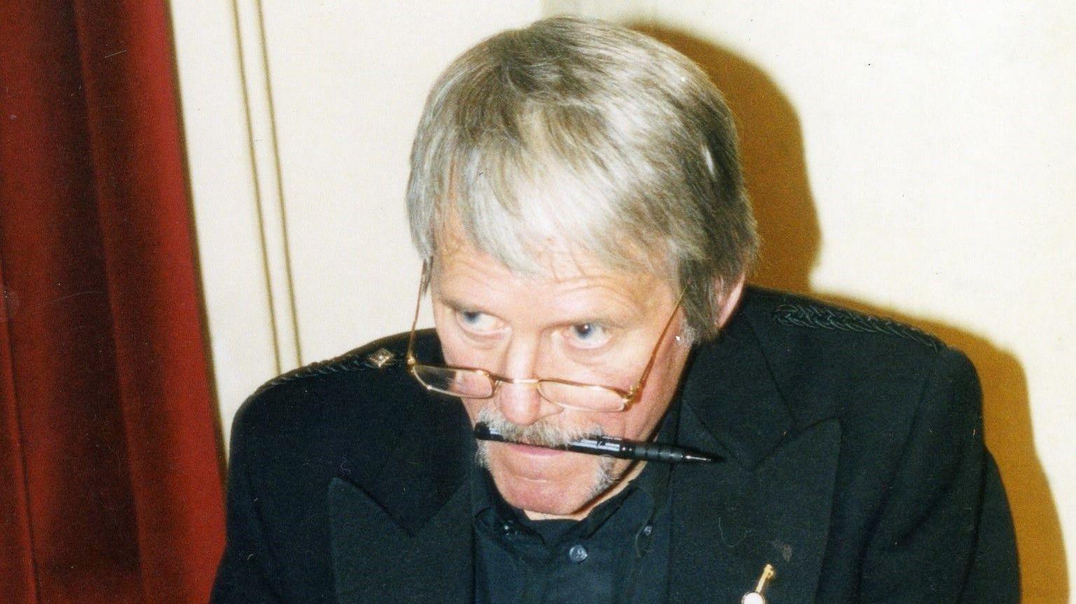 Malky McCormick in black jacket and shirt with a black felt tip pen in his mouth, looking up and to the left over a pair of gold-rimmed reading glasses. He has short, white hair and blue eyes.