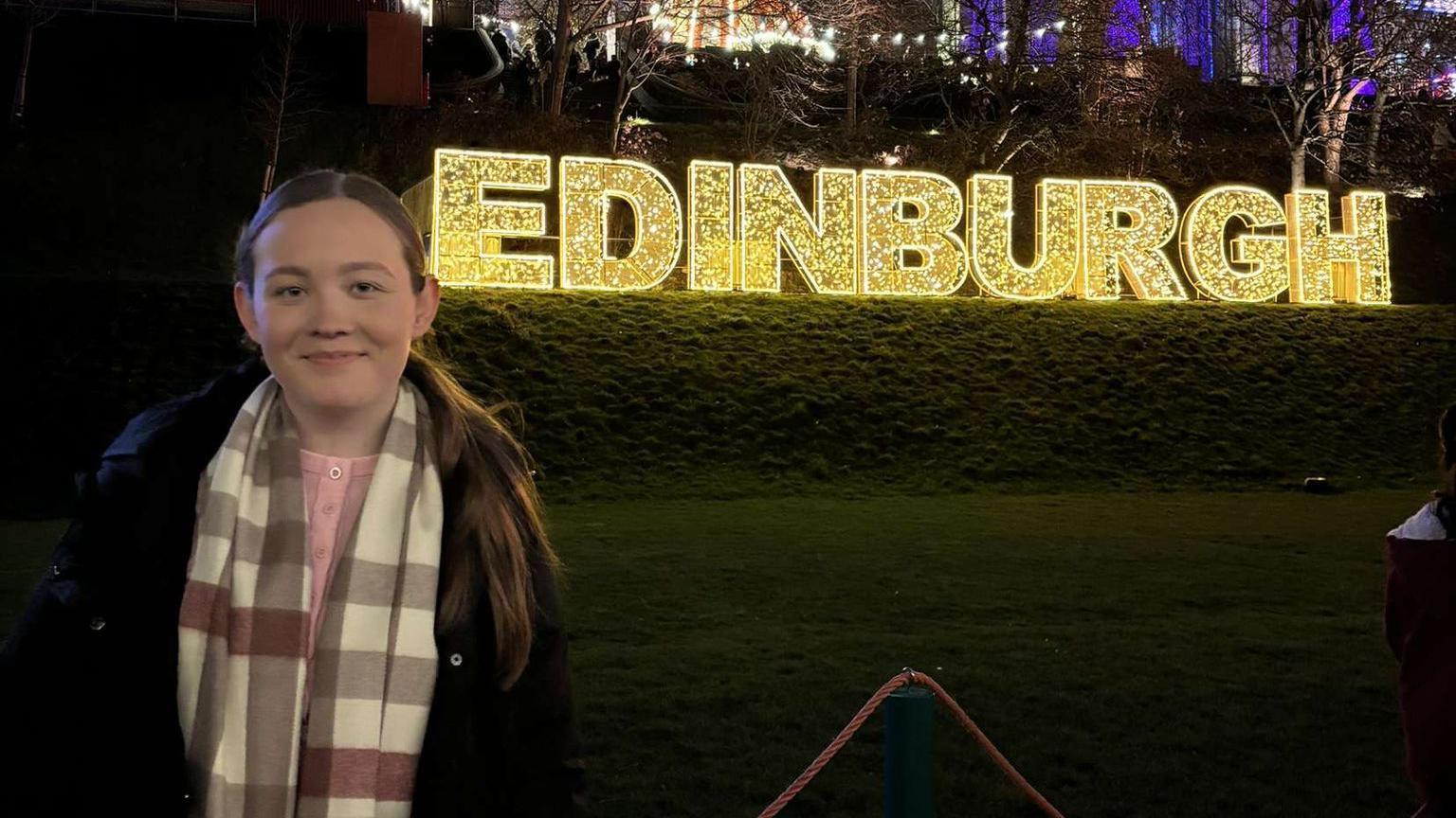 Rebecca is standing on the left smiling at the photo. She is wearing a coat and a scarf. She is standing by the word Edinburgh which is lit up. The photo was taken at night.