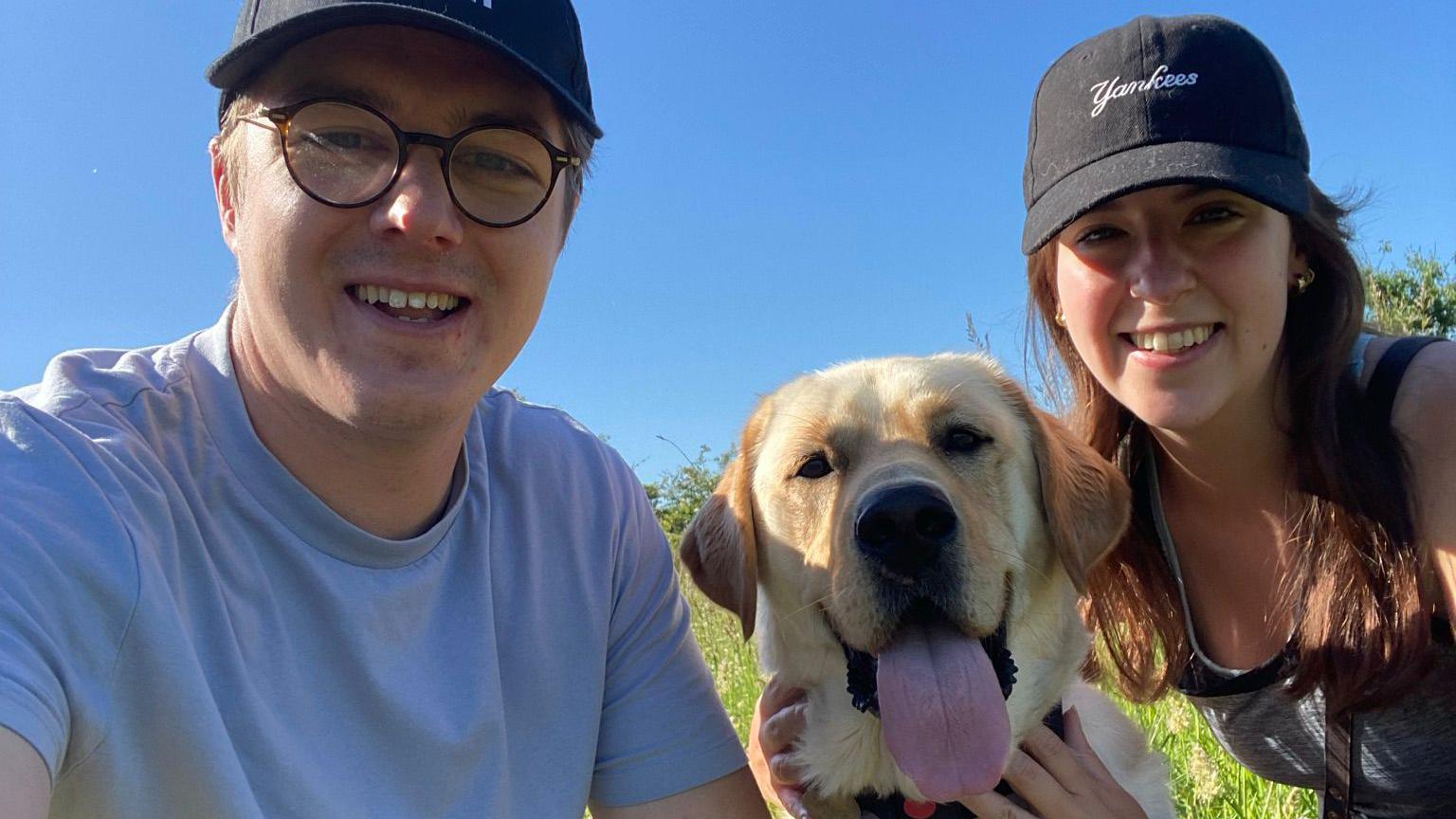 Adam Drysdale and Elle Manners pictured with a blonde labrador. Adam is wearing glasses and a cap and is smiling. Elle is also in a cap and smiling. The dog in the middle of them has its tongue out. 