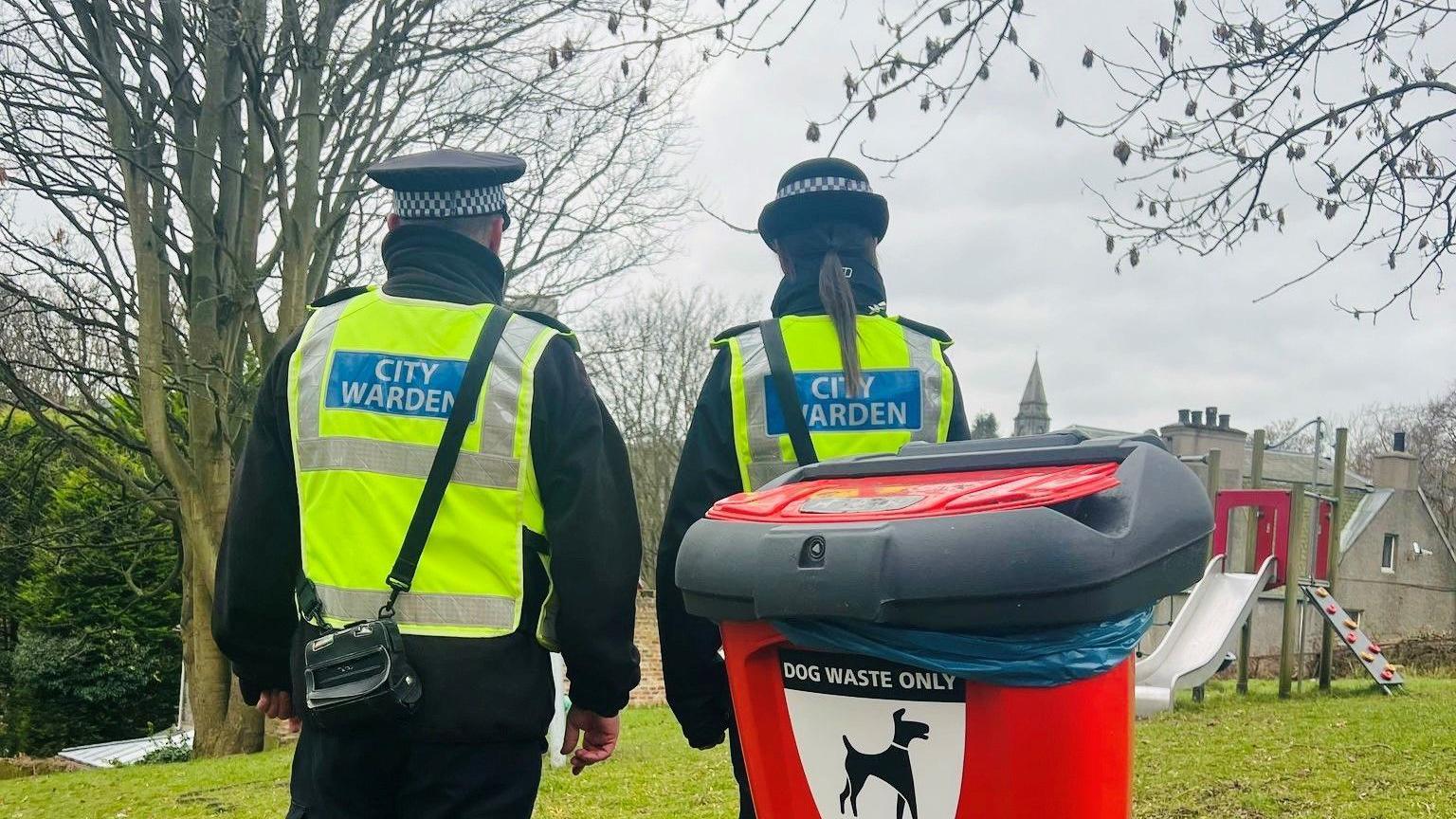 Two people with bright yellow 'City Warden' hi-vis jackets, standing on grass, behind a red dog waste bin.