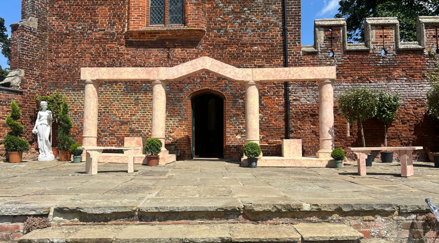 The flagstone stage has steps at the front and is a sandy colour. Behind it is are the red bricked walls which match that of Buckden Tower. There is some set on the stage which includes a white statue, green bushes and small trees. In the centre of the stage there is a door. Around this are four pillars - made to look like marble - and two matching benches at either side.