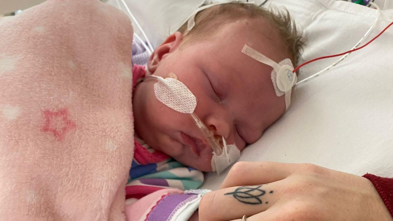 Newborn Arabella in a hospital bed with medical tape holding an oxygen tube on her nose and a monitor pad on her forehead part of a purple and pink striped babygrow is visible but she is wrapped in a soft pink blanket with stars on it. A woman's hand is holding the baby's hand, it has a flower tattoo on one finger and a silver flower ring on the middle finger.