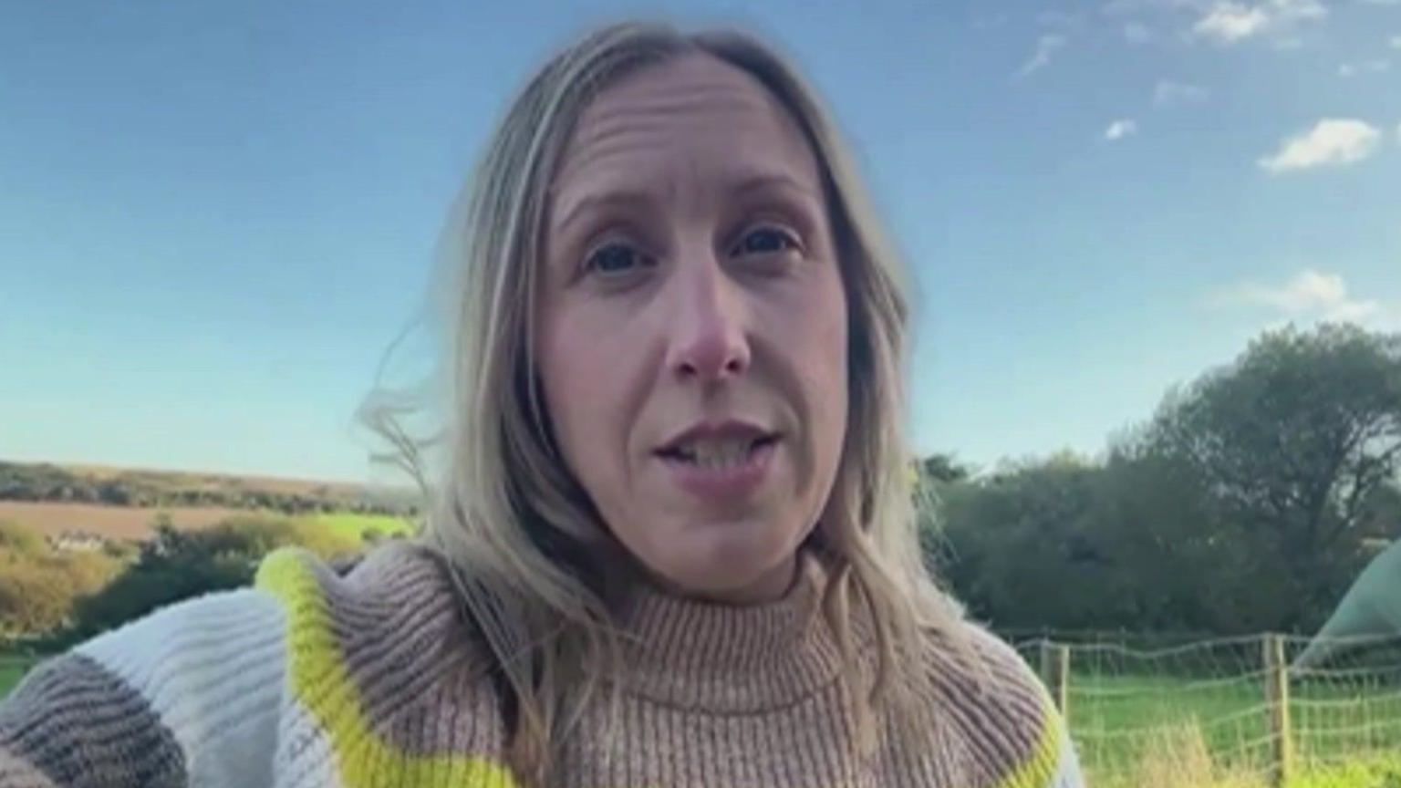 Claire Critchison is holding her phone up to do an interview, she is looking at the camera, has blonde mid length hair and wears a beige, yellow and white striped knit jumper. In the background are fields and farmland.