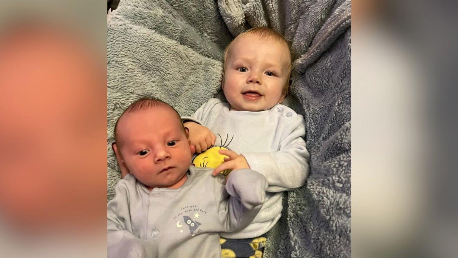 Archie and his baby brother Albie lying on a soft grey rug. Archie is smiling at the camera.