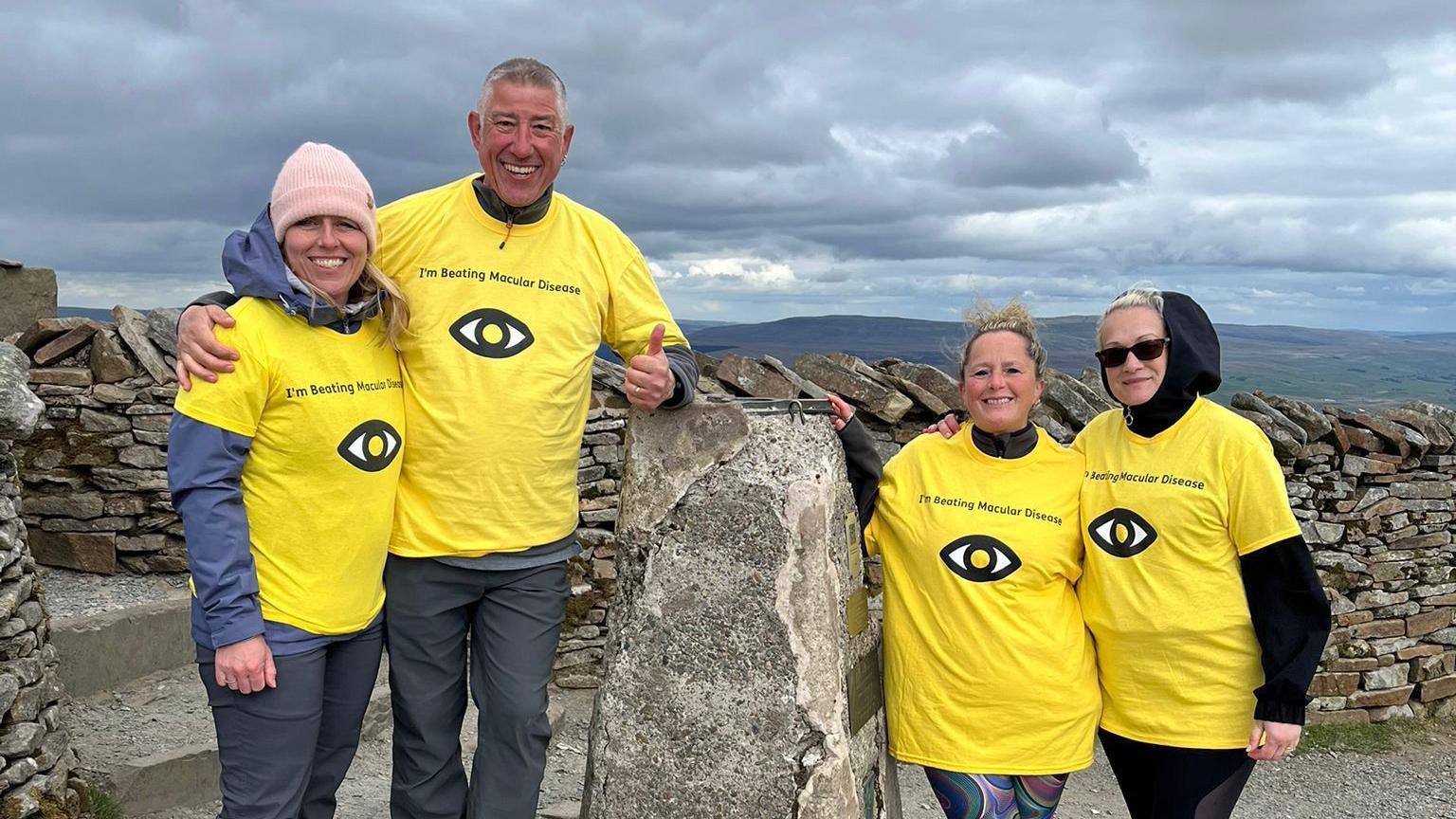 Mrs Blezard and some of the team stand at the top of a mountain. They all wear matching yellow T-shirts over the top of warmer clothes.