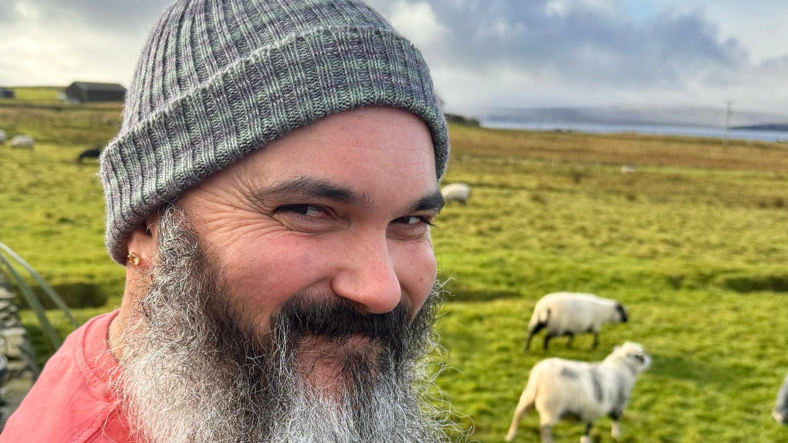 A man with long black and white beard wearing a grey knitted hat is standing in front of a green field in Shetland, with a few sheep visible behind him.